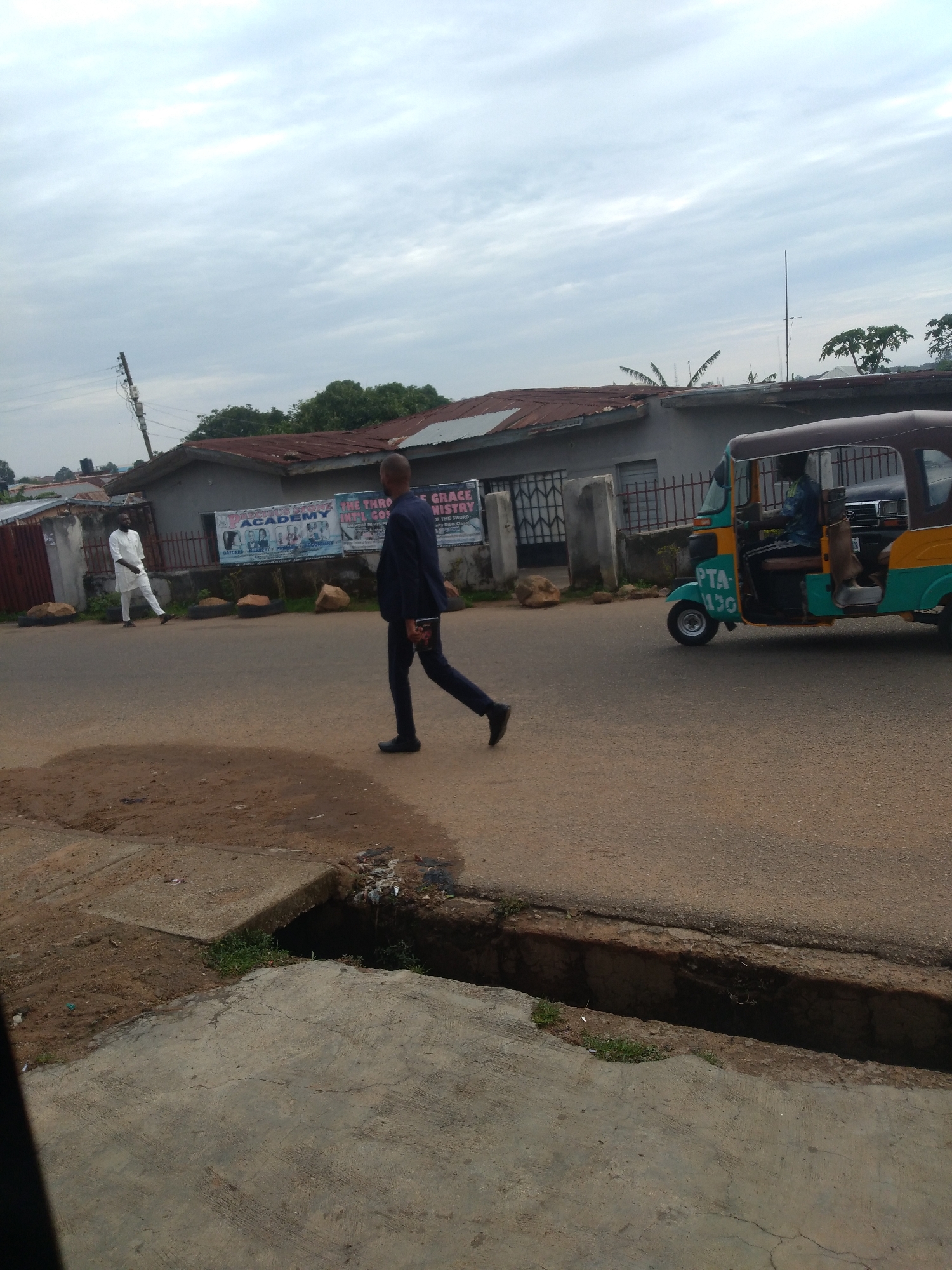 The image depicts an outdoor street scene in the city of Unknown, Unknown, under an overcast sky. A man in a dark suit walks from right to left across a paved road. On the left side of the road, another man in a white tunic and trousers stands near a metal fence. Behind the fence are one-story buildings with corrugated metal roofs. Two banners are displayed on the fence: one for "PRECIOUS STONE ACADEMY" and another for "THE THRONE OF GRACE MINISTRY." On the right side of the road, a teal and yellow three-wheeled vehicle, marked "PTA-130," is present with a driver and a visible passenger. In the immediate foreground, a concrete and dirt drainage ditch runs parallel to the road, containing some litter and sparse vegetation. Green trees are visible in the background above the buildings. A wooden utility pole with wires is visible on the far left.