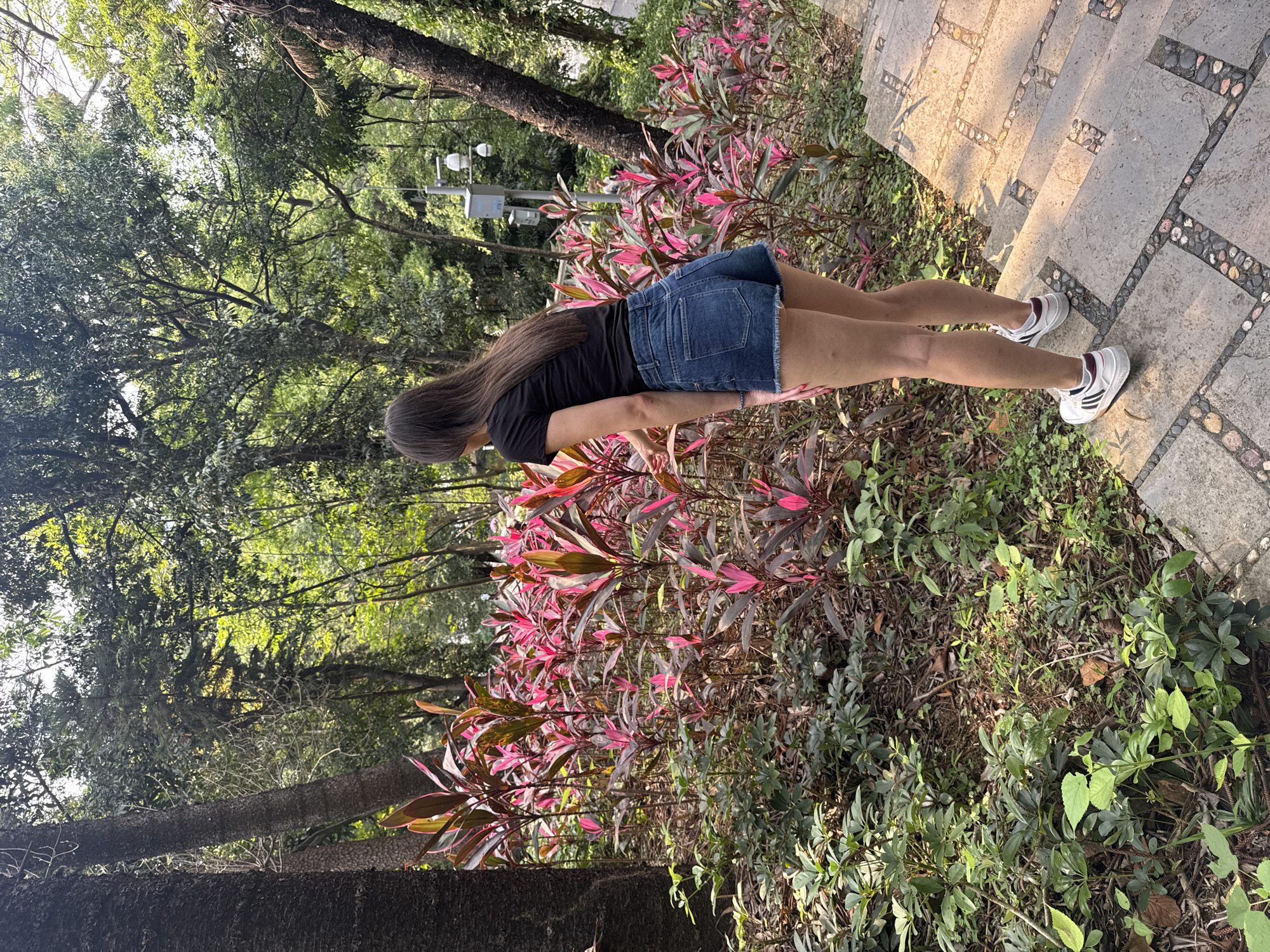 The image shows a young woman standing on a stone staircase with decorative edges in an outdoor, wooded area. The woman has long, dark hair and wears a black t-shirt, a denim skirt, and white sneakers with colored stripes. She appears to be near a planting bed with red and green foliage. In the background, there are trees and other greenery, suggesting a park or garden setting. The location is Yuexiu District, China.