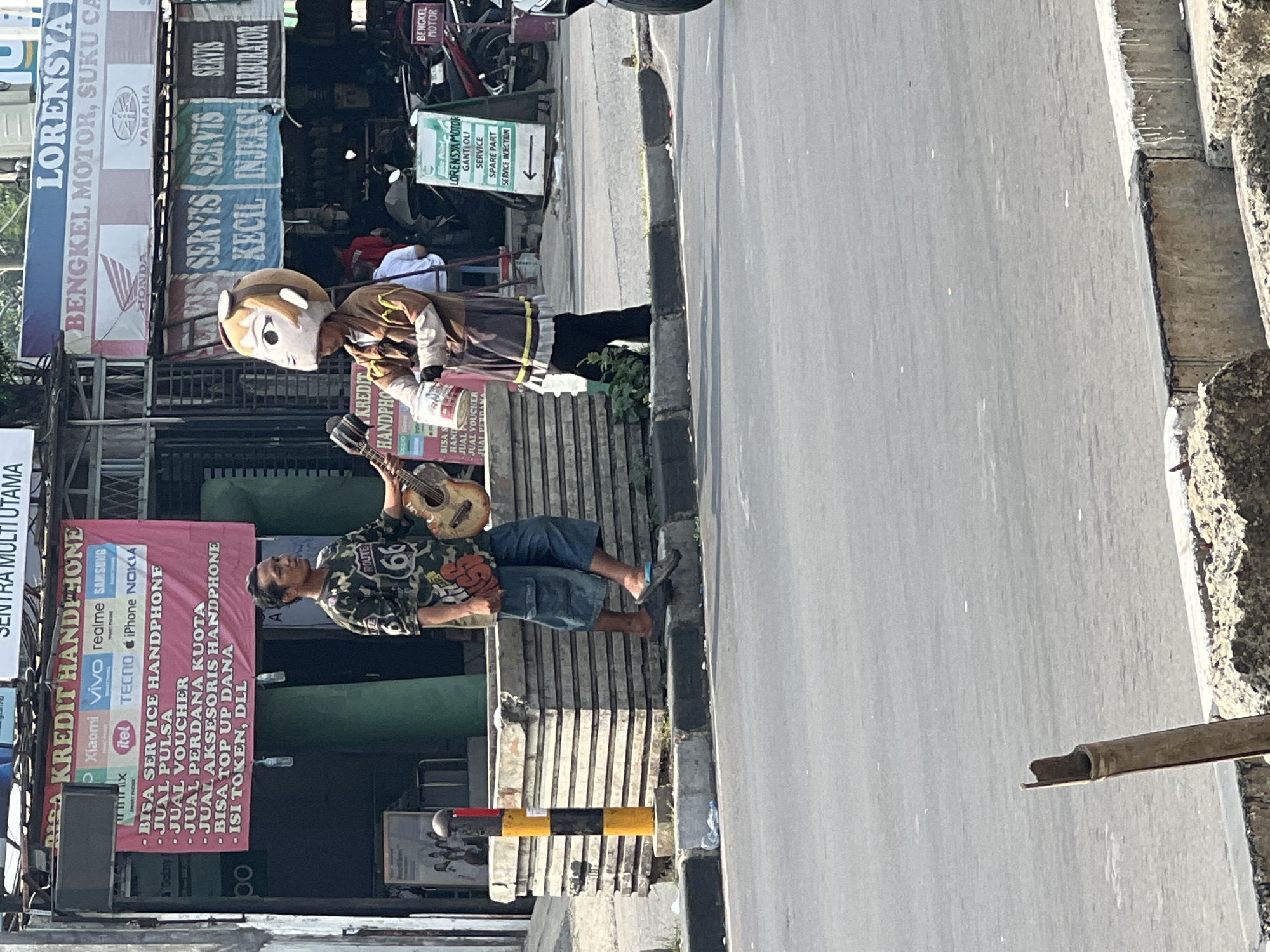 In Cibinong, Indonesia, on a sunlit day, a street performer wearing a large cartoon-like mask, possibly of a character with large eyes and blonde hair, stands by the roadside. This individual is dressed in a tan jacket with brown trim and dark pants, and holds a bucket in their right hand. Beside them, another person, dressed in a camouflage shirt and blue shorts, plays an acoustic guitar. Both individuals appear to be interacting, possibly performing for passersby. The scene is set in front of a storefront displaying a large red banner that advertises "KREDIT HANDPHONE" (phone credit) and various mobile phone brands. Adjacent to this, a shop selling motorcycle parts and services, "LORENSYA MOTOR", is visible. The backdrop includes parked motorcycles and various other signs, indicating a commercial area. The performers are positioned on a sidewalk next to a road with a concrete median barrier.