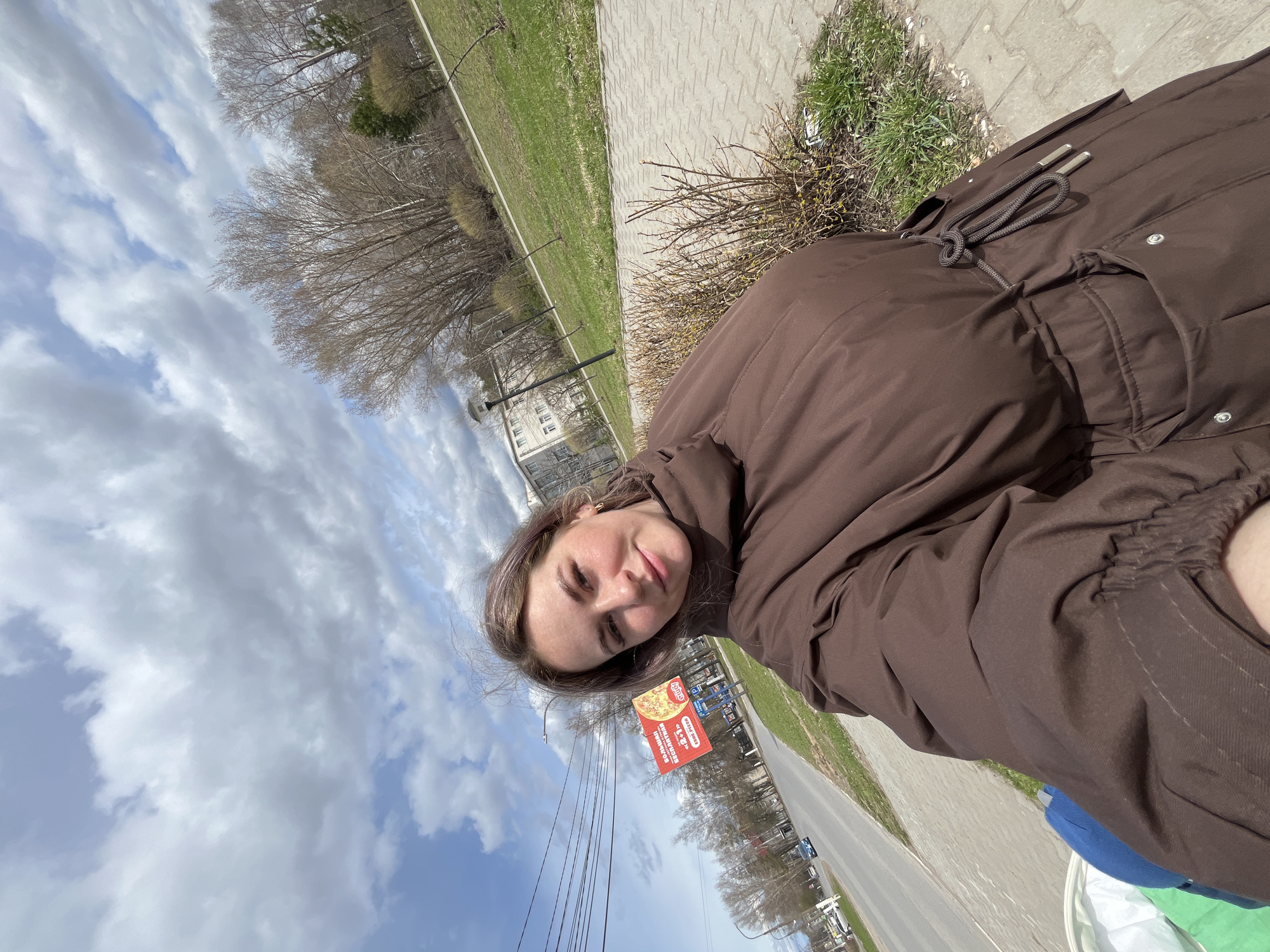 The image is a slightly tilted selfie taken by a young woman outdoors on a partially cloudy day. The woman, with long dark hair and a light complexion, looks directly at the camera with a gentle, faint smile. She is wearing a warm, brown puffer jacket or coat, featuring a drawstring waist with silver toggles.
The background depicts an urban or suburban landscape during what appears to be late winter or early spring. To the woman's left, a paved sidewalk runs alongside a strip of green grass and a scattering of bare trees and leafless bushes. Further back, a road is visible, lined with streetlights and power lines. In the distance, several multi-story buildings can be seen under a sky with both blue patches and substantial white cumulus clouds.
A prominent red and white billboard is visible behind the woman, advertising pizza. It features the English word "PIZZA" and Cyrillic text that translates to "Large Free," likely promoting a "Dodo Pizza" offer, indicated by a partially visible logo. The scene suggests a casual moment captured during daytime in an outdoor setting, consistent with the provided location context of Chaikovsky, Russia, due to the Cyrillic text and architectural cues.