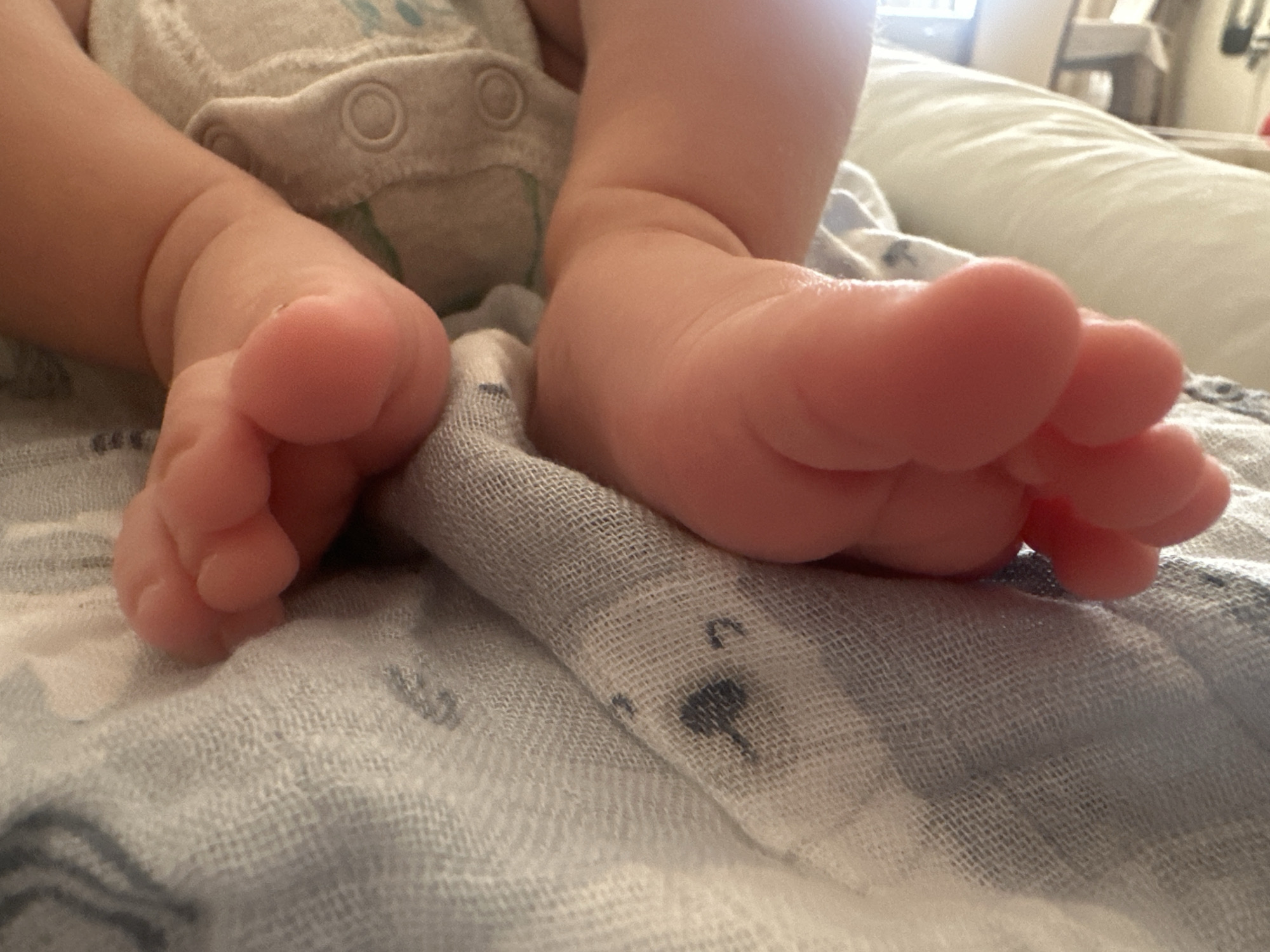 This is a close-up, eye-level shot of a baby's feet resting on a light blue and white patterned muslin cloth. The baby is wearing a grey and white diaper. The focus is on the baby's feet, with the toes and soles clearly visible. The background is slightly blurred, showing a white, cushioned surface and a hint of a light-colored room. The lighting suggests an indoor setting. The location in Masazir, Azerbaijan, is not discernible from the visual information provided in the image.