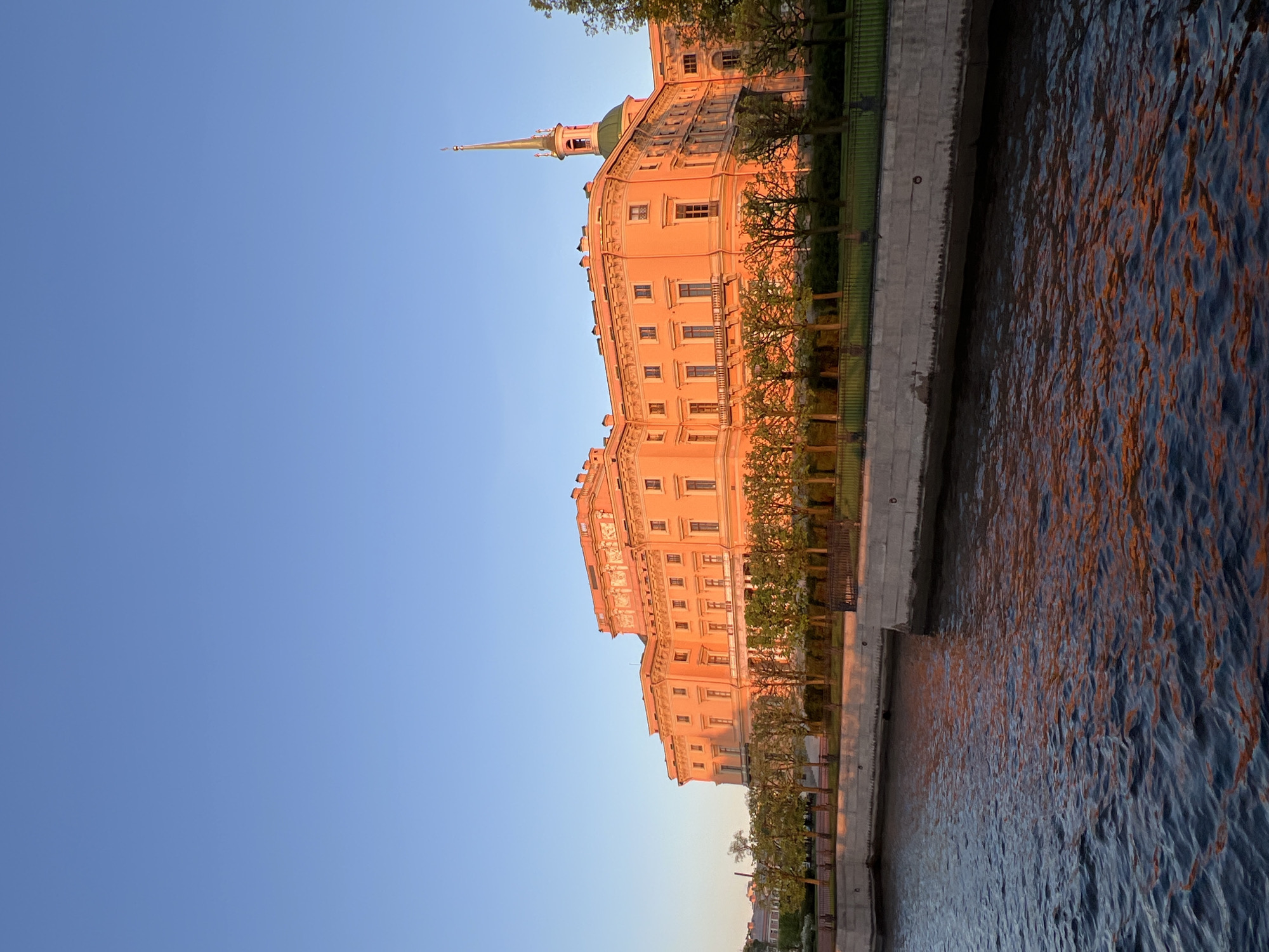The image shows the Saint Michael's Castle in Saint Petersburg, Russia, from a low angle. The castle is a peach-colored building with a prominent spire. It is situated next to a body of water, likely a canal, where the orange light of the setting sun is reflected on the water surface. Along the embankment, there is a row of trees planted. The sky is clear and blue.