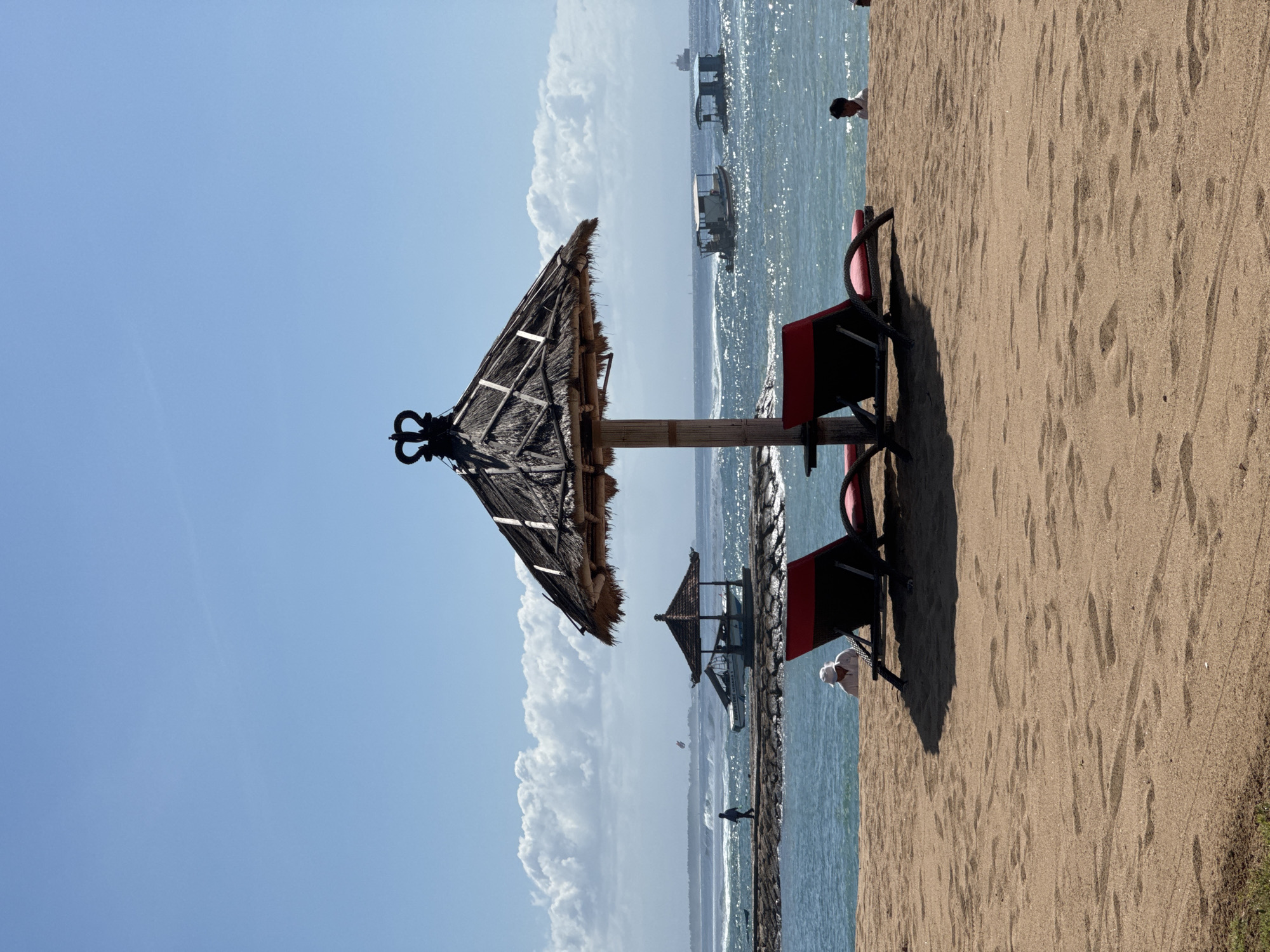 The image captures a beach scene in Kuta Selatan, Indonesia. A thatched umbrella stands prominently, with two dark-colored lounge chairs adjacent to it. The sand shows many footprints. The water shimmers in the sunlight, and several boats are visible in the distance. Further into the sea is a rock-lined pier and a small gazebo on the pier. A few people are walking on the beach and the pier. White, fluffy clouds populate the blue sky.