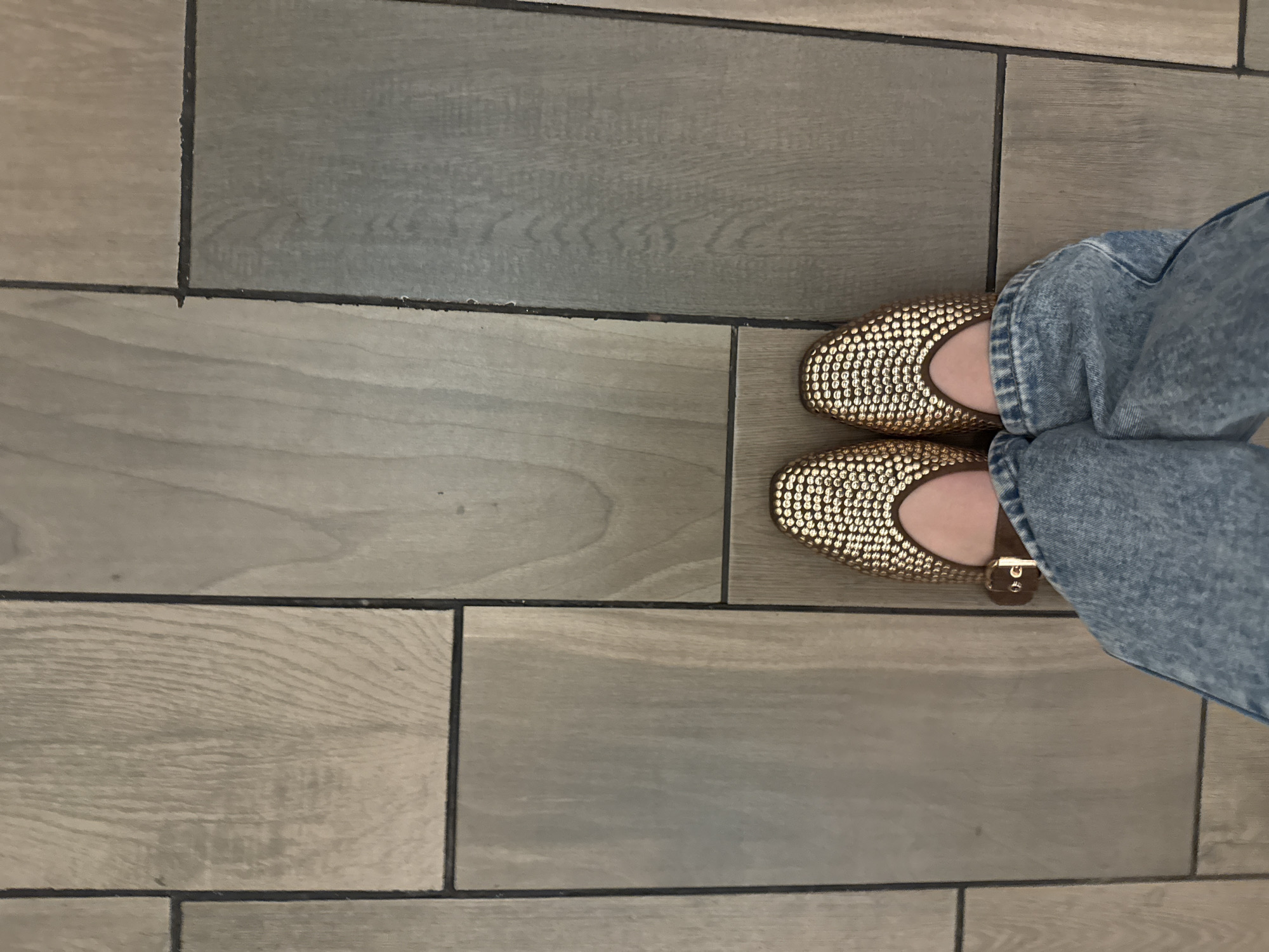 The image depicts a person standing on a tiled floor, wearing blue jeans and a pair of gold-colored, studded ballet flats. The tiles appear to be wood-look, with a rectangular pattern and varying shades of gray and brown. The floor is clean and well-maintained. There is no other information in the image to determine if the location is in Mexico City, Mexico.