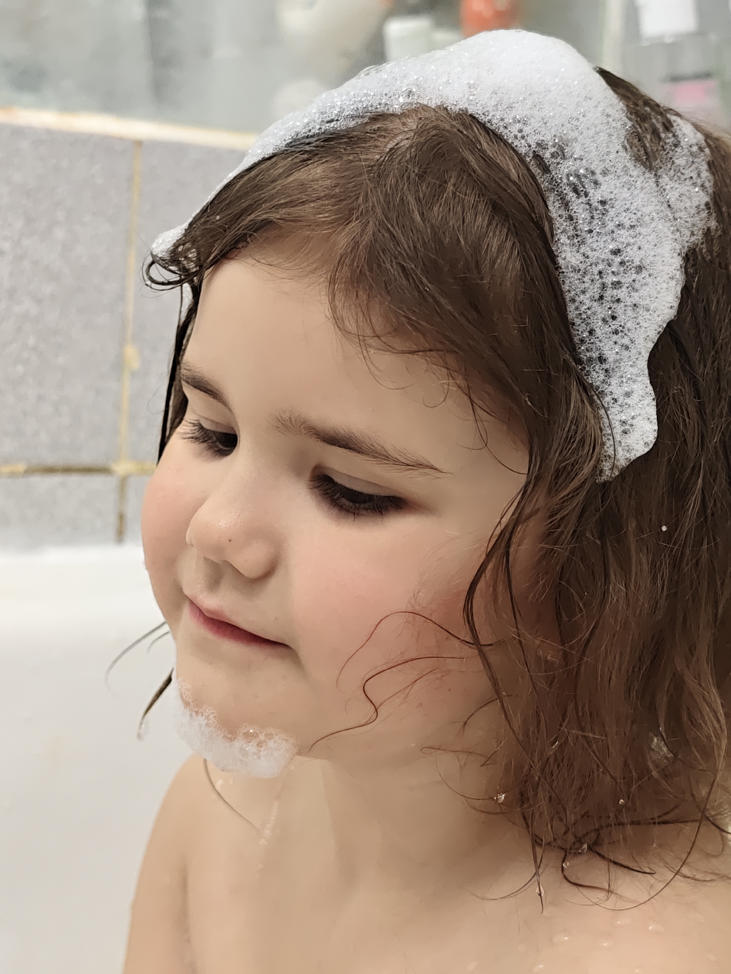 In a close-up, vertical shot, a young child with brown, wet hair is depicted in a bathtub in Chaikovsky, Russia. The child's profile is visible, facing towards the left side of the frame, with her gaze directed downwards. Her expression is gentle and slightly contemplative, with a subtle smile playing on her lips. Her cheeks appear rosy.
A significant amount of white foam or bubbles, likely from soap or shampoo, rests on top of her head, forming a band along her hairline, and also on her chin. Her hair is damp and shows slight waves and curls, with individual strands clinging to her face and shoulders. Water droplets are visible on her wet hair.
The background consists of light-colored, possibly tiled, bathroom walls, partially blurred, and the white rim of the bathtub. The scene suggests the child is in the middle of a bath or having her hair washed, capturing a moment of quiet contentment or engagement with the water and bubbles. No text is visible in the image.