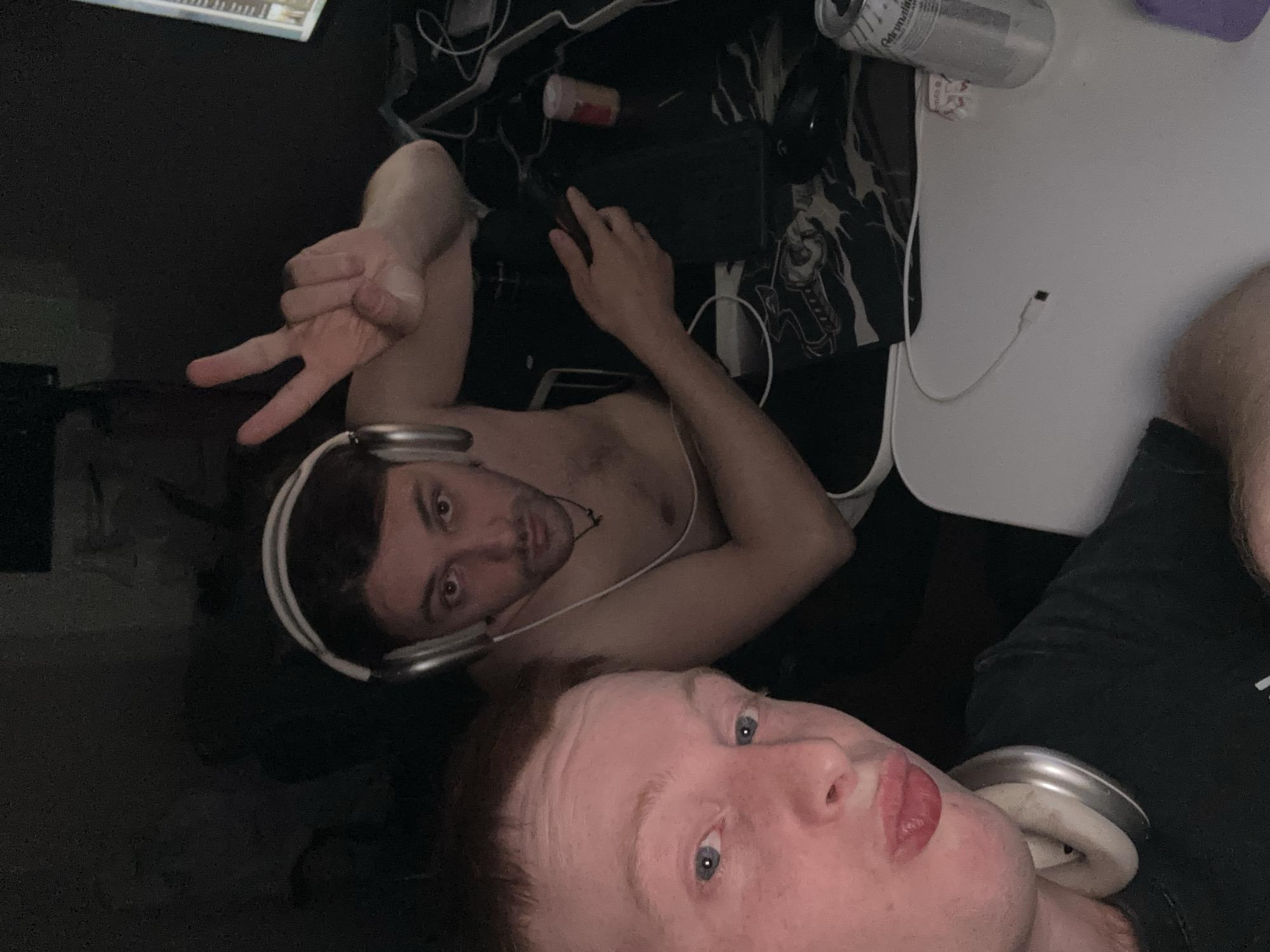 Two men are visible in the image. One man is lying on his back wearing headphones and shirtless, holding up a hand gesture. The other man is positioned closer to the foreground, wearing headphones and a black shirt, and looking at the camera with pursed lips. The setting appears to be indoors, with a computer keyboard, mouse, and various objects scattered around, including a can of "Adrenaline Rush". The image does not contain location information for Ореховка, Russia.