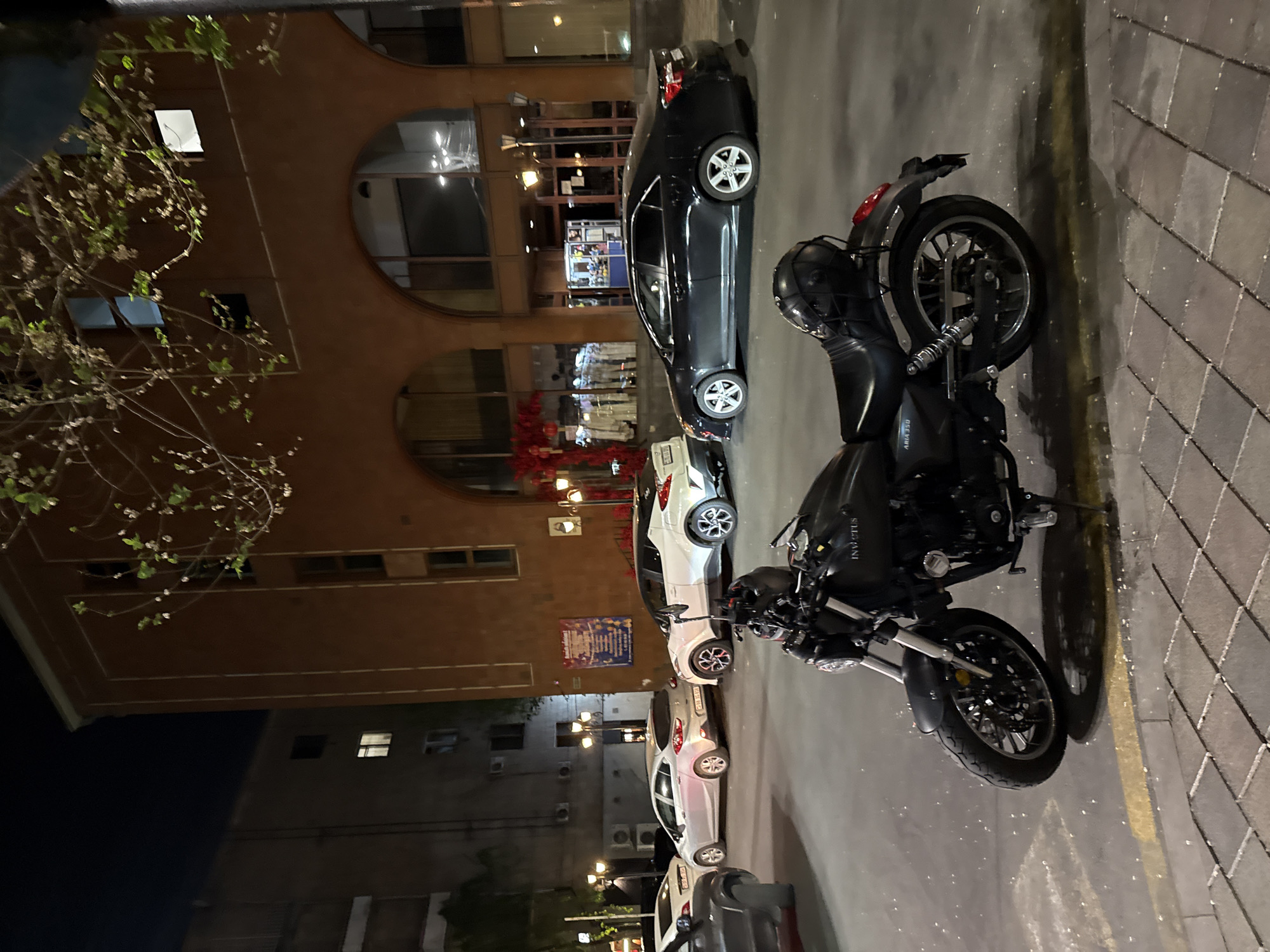 The image depicts a street scene in Yerevan, Armenia at night. In the foreground, a black motorcycle with "INVUCTUS" lettering is parked on a grey paved surface. A dark sedan is parked behind the motorcycle. Further back, several other vehicles, predominantly white, are parked along the street.
A multi-story brown building occupies a significant portion of the background. The building's architecture includes rounded archways and illuminated windows, suggesting commercial or residential units. Streetlights illuminate the scene, and some of the shop windows are lit. A tree is visible on the left side of the image. Another building with smaller windows is visible in the distance on the left.
The overall atmosphere is that of an urban environment at night.