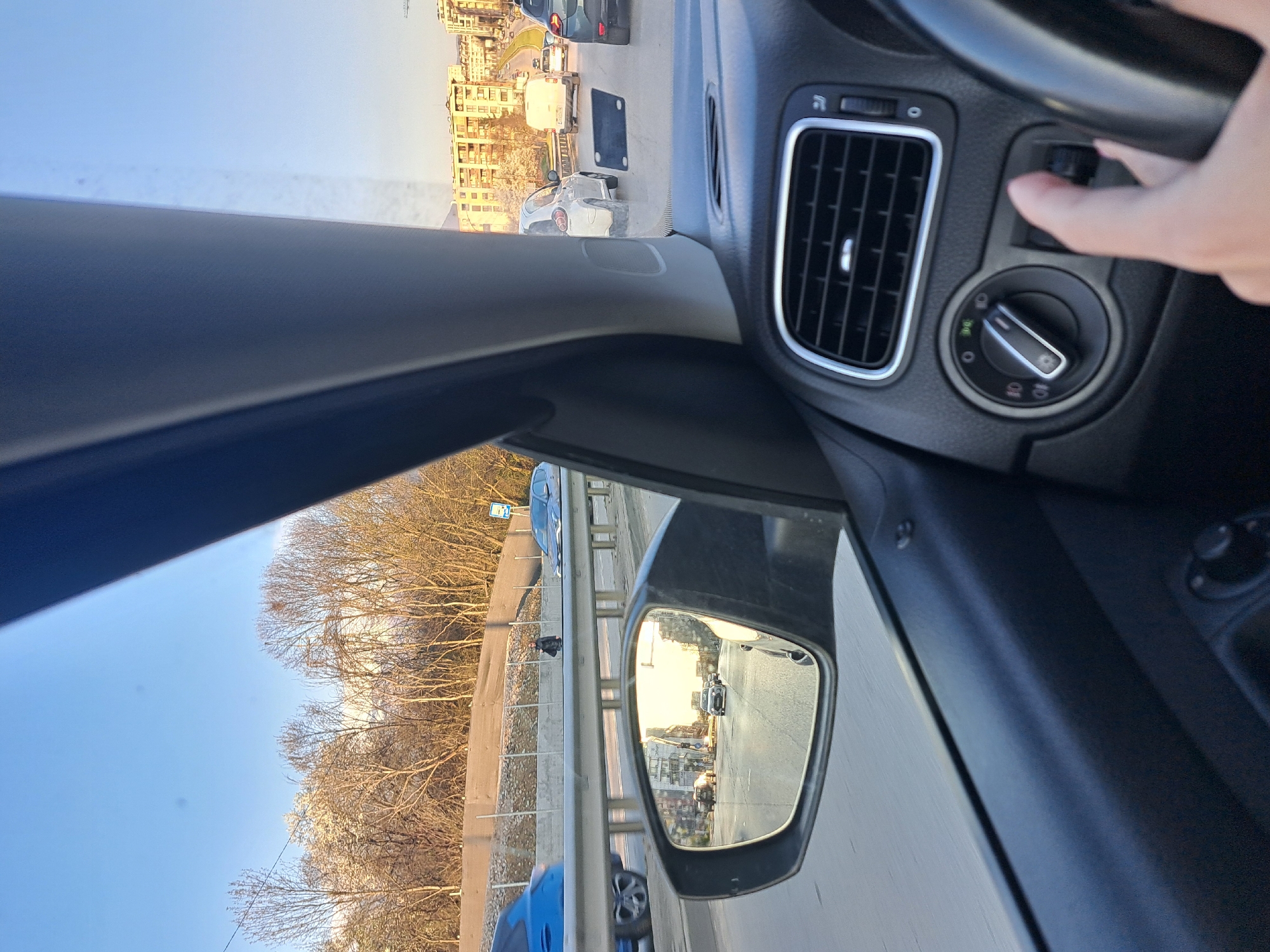 The image depicts a view from inside a car, likely in Sofia, Bulgaria, due to the architecture visible in the background. The driver's hand is visible, operating a knob on the dashboard. The car is in traffic, with other vehicles seen ahead and in the side mirror. The side view mirrors capture an image of a multi-lane highway, separated by a metal guardrail. Beyond the rail are bare trees. Buildings are visible in the distance. The weather is sunny and clear. The scene is a typical traffic situation on an urban or suburban highway.
