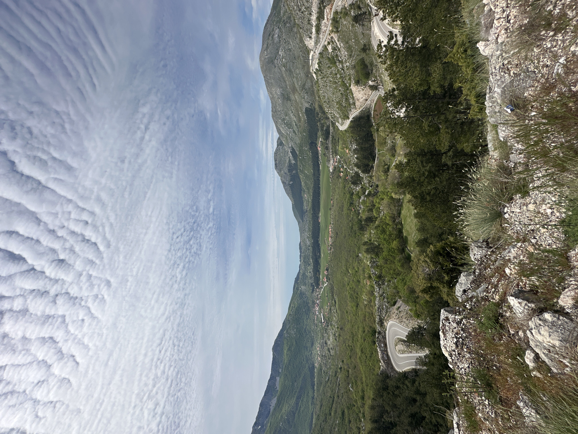 A landscape photograph depicts a mountainous region in Bukovica, Montenegro. The sky, filled with striated clouds, takes up approximately half of the left side of the image. A steep, rocky mountain dominates the right side, covered in sparse vegetation and glimpses of a winding road. Below the mountain is a green valley with scattered vegetation and a few visible buildings. The viewpoint is from a rocky outcrop with small shrubs and grasses.