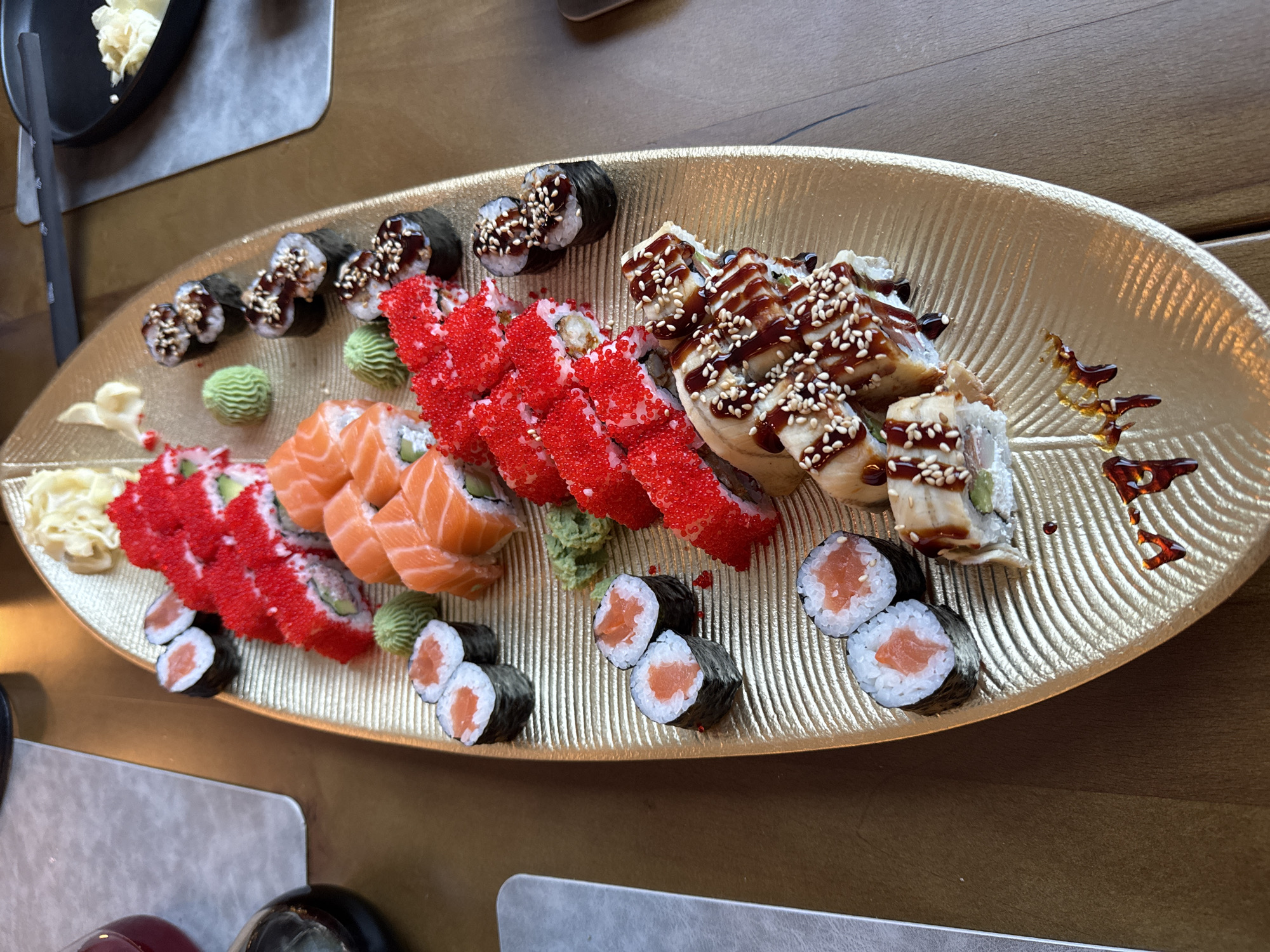 A high-angle, eye-level shot captures a large, ornate, gold-colored platter heavily laden with various sushi and maki rolls, presented on a dark wooden table in Budva, Montenegro. The gold platter, shaped like an elongated leaf with a textured, ribbed surface, serves as the central focal point.
Arranged on the platter are multiple types of sushi. Towards the upper left, a line of maki rolls is topped with vibrant red tobiko or masago. Adjacent to these, a row of orange salmon nigiri or similar rolls with raw salmon draped over rice is visible. Another section displays traditional maki rolls, dark with seaweed, drizzled with a dark glaze and sprinkled with white sesame seeds. Further down, pairs of smaller maki rolls with salmon are arranged. The bottom right segment of the platter features larger, more elaborately constructed rolls, likely featuring eel or similar fillings, generously coated in a dark, glossy sauce and topped with white sesame seeds.
Accompanying the sushi are several garnishes: a mound of pale yellow pickled ginger (gari) is positioned at the top right of the platter, and several small, green, sculpted mounds of wasabi are distributed among the sushi arrangements. Dark, viscous sauce drizzles are decoratively applied to the lower section of the gold platter.
The immediate environment includes a dark wooden table. To the left, a light-colored placemat, a dark glass bottle, and a clear glass carafe containing a dark red liquid are partially visible. To the right, another light-colored placemat and a black, shallow pan holding a small quantity of light-colored food are present in the background. No people are visible within the frame. The lighting is soft, creating minimal shadows and highlighting the metallic sheen of the platter.