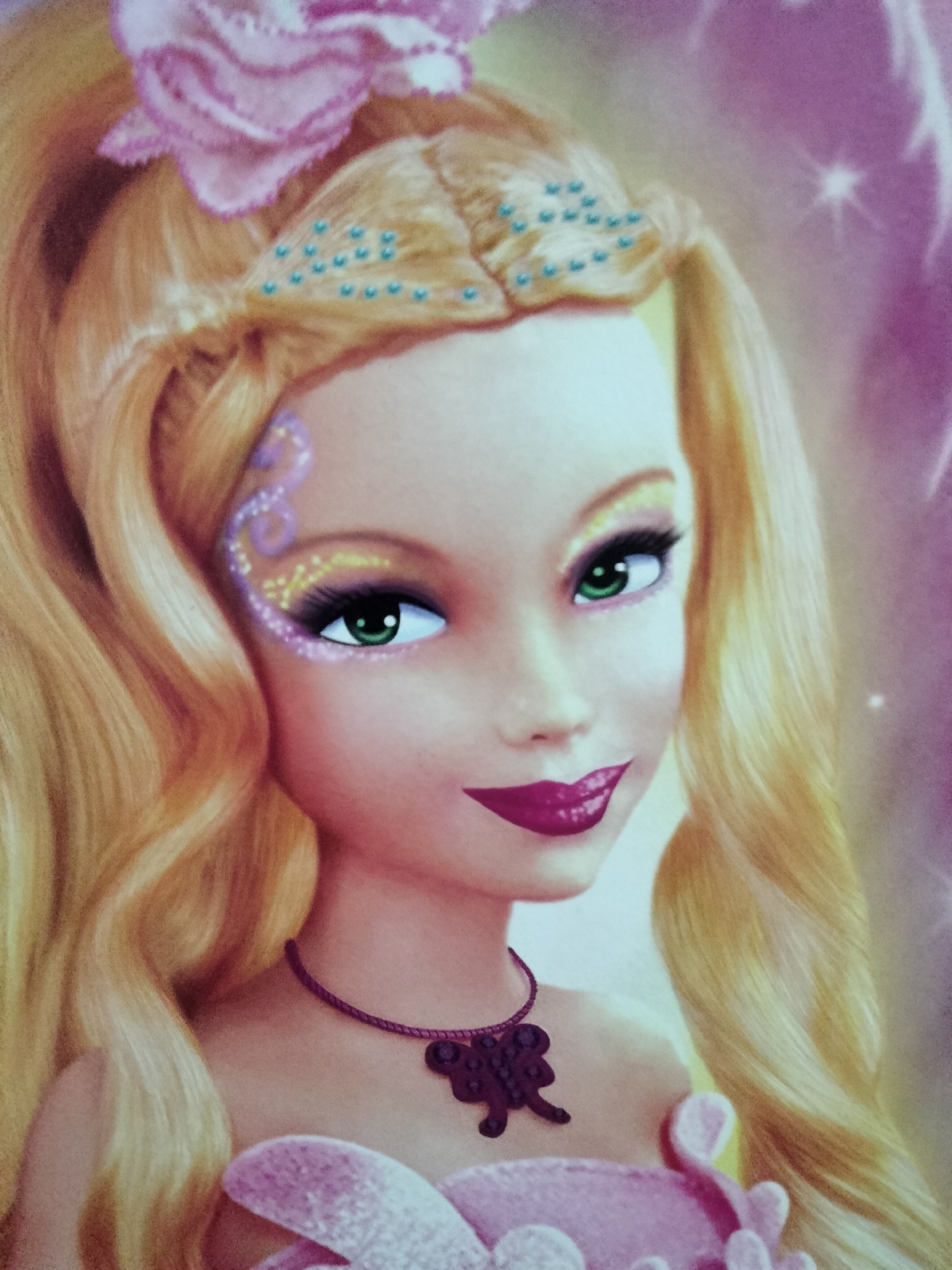 The image is a close-up portrait of a doll with long, blonde, wavy hair. She has green eyes, bright pink lipstick, and is wearing a pink dress and a purple necklace with a butterfly pendant. Her hair is adorned with a large pink flower and blue beads. The background is a soft, blurred pink and purple gradient with some sparkling light effects. There is no indication of the city of Payagaboh, Indonesia, or any specific location within it.