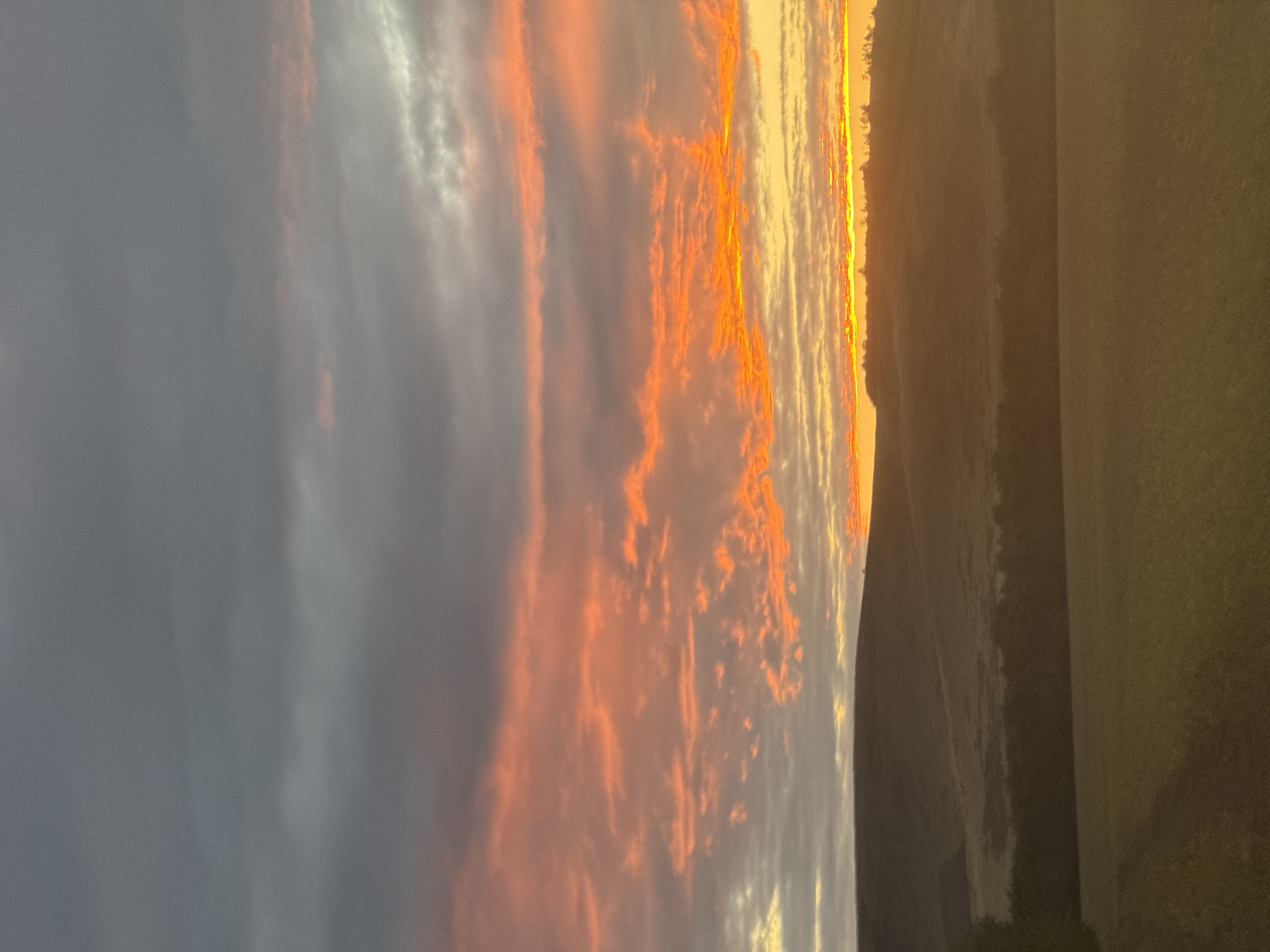 The image depicts a sunset scene over a rural landscape, possibly in the vicinity of Vasil'evka, Russia. The sky is dominated by dramatic clouds, with the upper portion being a dark, stormy gray and the lower portion illuminated by vibrant hues of orange and yellow from the setting sun. Wisps of pink and red streak across the horizon. Below the sky, rolling hills are silhouetted against the bright sky, their contours softened by the fading light. A line of trees runs along the crest of the hills. In the foreground, a field is visible, its surface a muted green and brown, suggesting cultivated land or grassland. The overall atmosphere is serene and picturesque, capturing the natural beauty of a rural sunset. No people or animals are visible in the frame.