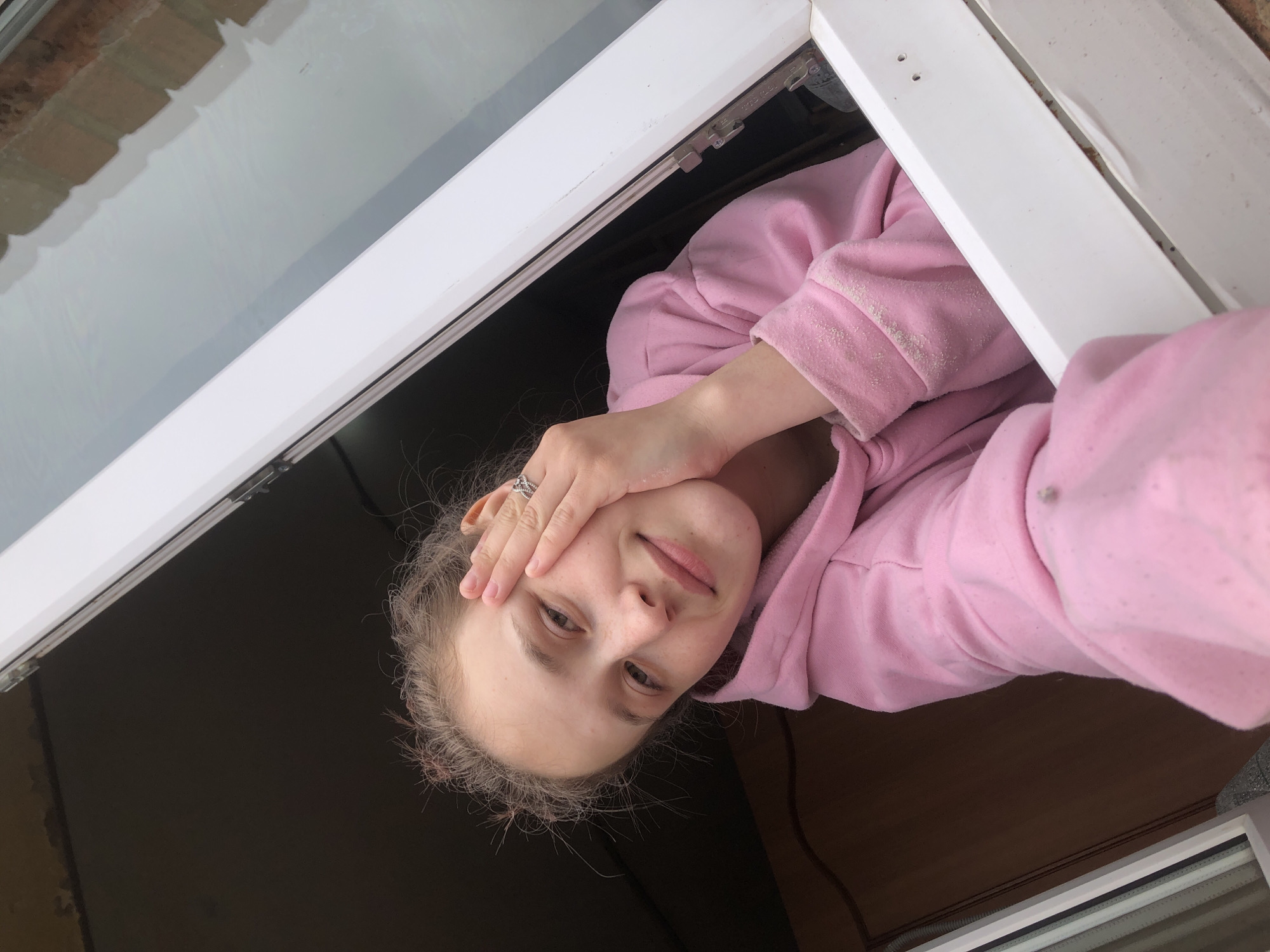 A woman wearing a pink hoodie peers into the camera from a window. She has fair skin, blonde hair, and is touching her forehead with her left hand. A silver ring is visible on her left ring finger. The window frame is white. The location of Фоки, Russia cannot be confirmed as there are no visible details or location data to determine the location.