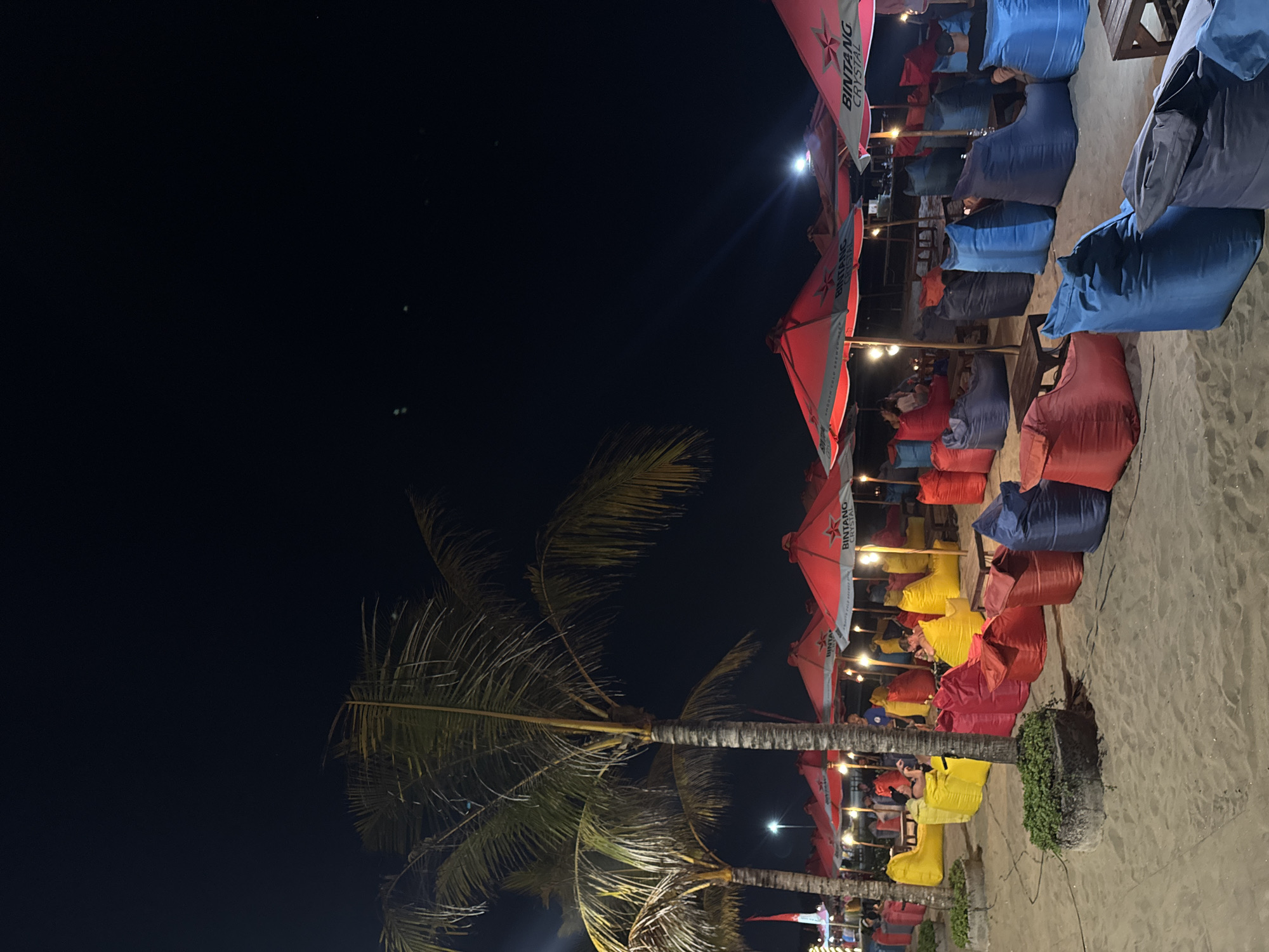 The image depicts an outdoor seating area on a sandy beach at night, likely in Kuta, Indonesia, given the presence of red Bintang beer umbrellas. The scene is lit by artificial lights, including the overhead lights of the umbrellas and some ambient light sources. Rows of colorful beanbag chairs in red, yellow, blue, and purple are arranged on the sand, forming seating arrangements for patrons. Several patrons are seated in the beanbags, engaged in conversations and enjoying the evening. Two palm trees with their fronds illuminated stand prominently on the left side of the frame, adding to the tropical ambiance. In the background, more seating areas and umbrellas are visible, suggesting a bustling beachfront establishment. The night sky is dark, with a few faint lights visible in the distance, possibly from other establishments or the town. The overall atmosphere is relaxed and social, indicative of a popular nighttime leisure spot.