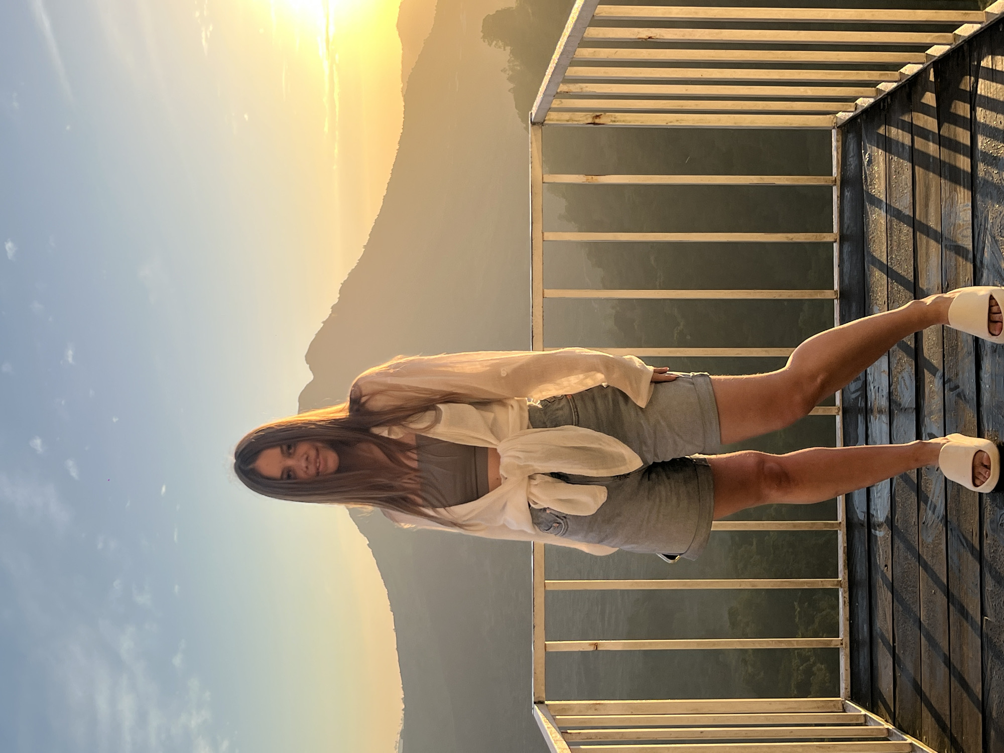 A young woman stands on a wooden balcony with a white railing, facing the camera. She is wearing a grey crop top, a white unbuttoned shirt tied at the waist, and denim shorts. She has long brown hair and is looking at the camera with a smile. The background features a mountain range bathed in the warm, golden light of a sunset, with a clear sky above. The location is identified as Kintamani, Indonesia.