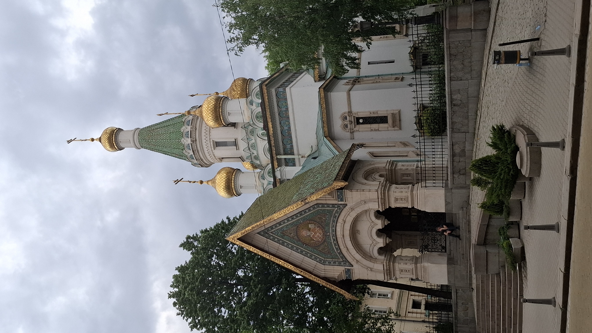 The image features the Church of St. Nicholas the Miracle-Maker, located in Sofia, Bulgaria. The church exhibits distinctive architectural features, including golden domes and a green tiled roof. The facade is white with ornamental details.
A person is sitting on the steps of the church, near its entrance. The area around the church includes a stone wall, black iron fencing, and paved walkways. There is also a small tree or shrub in a planter and street fixtures nearby. The sky is overcast.