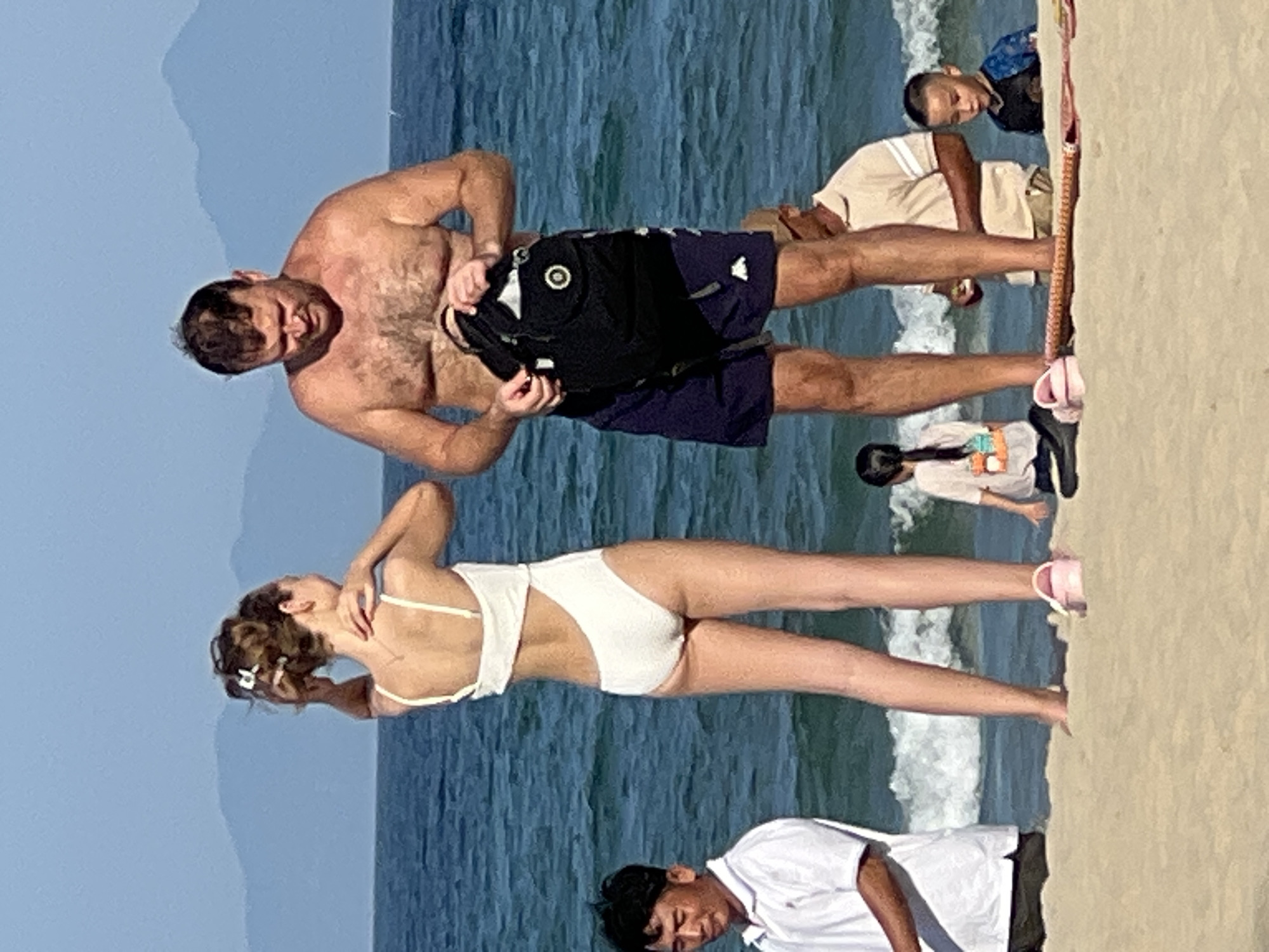 This image captures a scene at the beach in Đà Nẵng, Vietnam, likely during the daytime given the bright sunlight and clear blue sky. The ocean, with its gentle waves, forms the backdrop, suggesting warm weather suitable for beach activities.
The foreground is dominated by several people. A man, shirtless and wearing dark swim shorts, is visible in the upper left quadrant. He appears to be holding a black backpack. To his right, a woman in a white one-piece swimsuit stands with her back to the camera, her arms raised as if stretching or posing. Her hair is tied up, and she's wearing white sandals. In the lower right, a person wearing a white t-shirt is partially visible, their head tilted down. Further in the background, other individuals are seen, including a man in a light-colored shirt and shorts, and a child or younger person in a colorful top and shorts, walking on the sand.
The overall atmosphere seems relaxed and casual, indicative of a day spent at the beach. The clear weather and the presence of people enjoying the seaside suggest a pleasant day. No text is readily visible in the image.