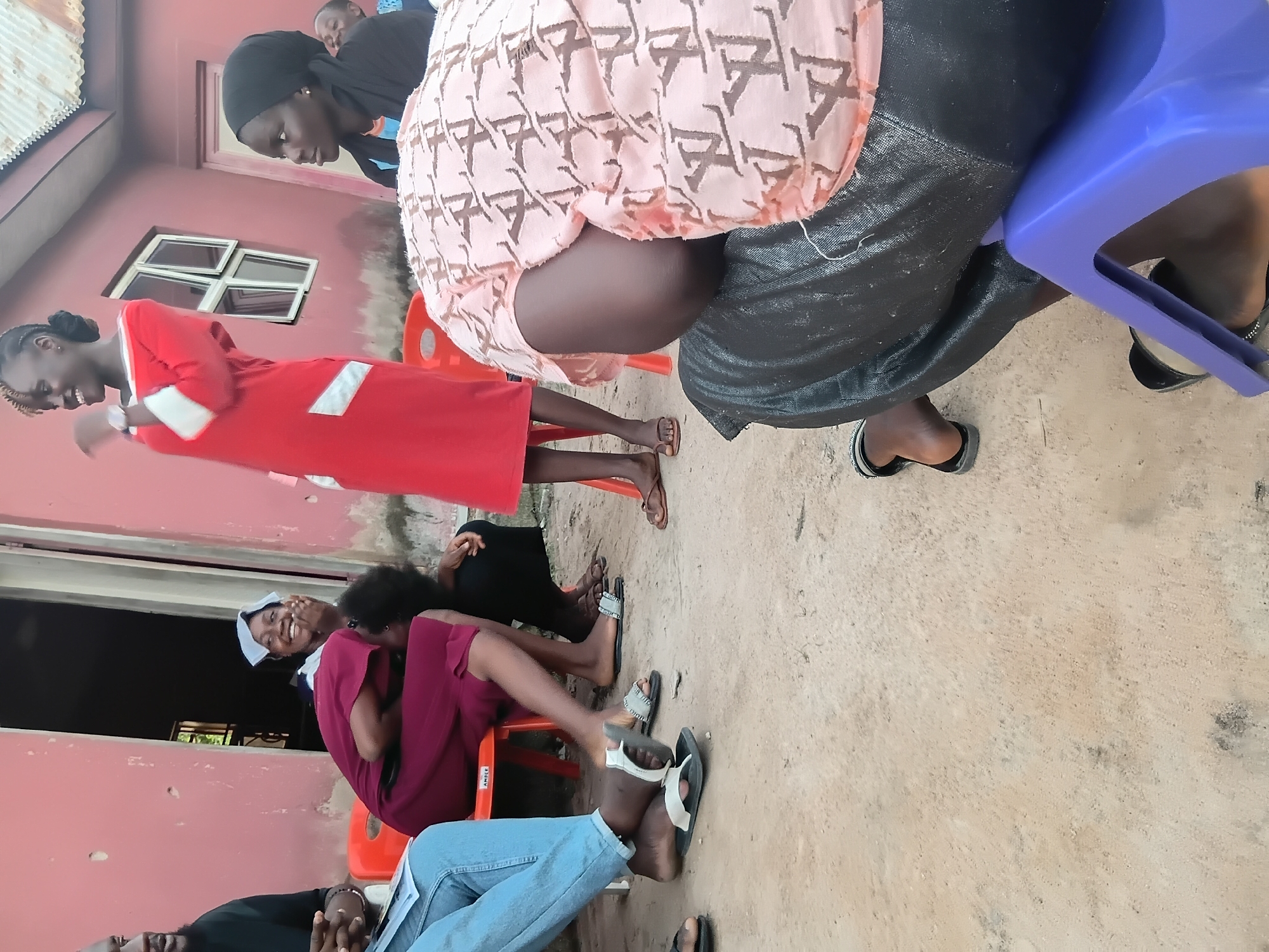 In Jalingo, Nigeria, an outdoor gathering takes place in front of a pink building with an open doorway and a corrugated metal roof. Several individuals are seated and standing. A woman in a red dress with white accents stands and gestures. Another woman in a dark headscarf looks towards the standing woman. A person in a burgundy dress is seated, laughing. Two individuals in the foreground are partially visible, one wearing jeans and the other a pink patterned top over dark pants. The ground is dirt and gravel. Orange and blue plastic chairs are present. The scene appears to be informal and social.