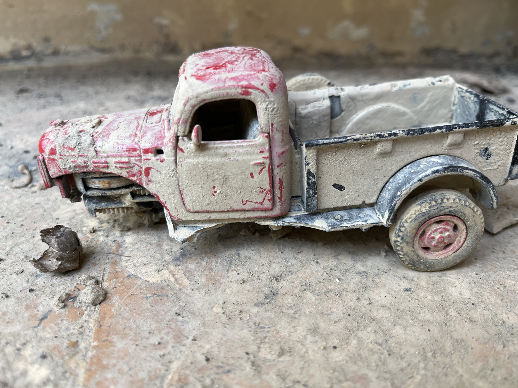 An overhead, close-up shot shows a dusty, red and tan toy pickup truck positioned on a textured, concrete surface. The truck is covered in a layer of light-colored dust and dirt, obscuring much of its original paint and detail. The front of the truck appears damaged, with visible metallic components and a tire missing. A small, dark, crumpled object lies on the ground to the left of the truck. The background is blurred, showing a beige, textured wall. The scene appears to be outdoors, possibly in an abandoned or neglected area. No people are visible.