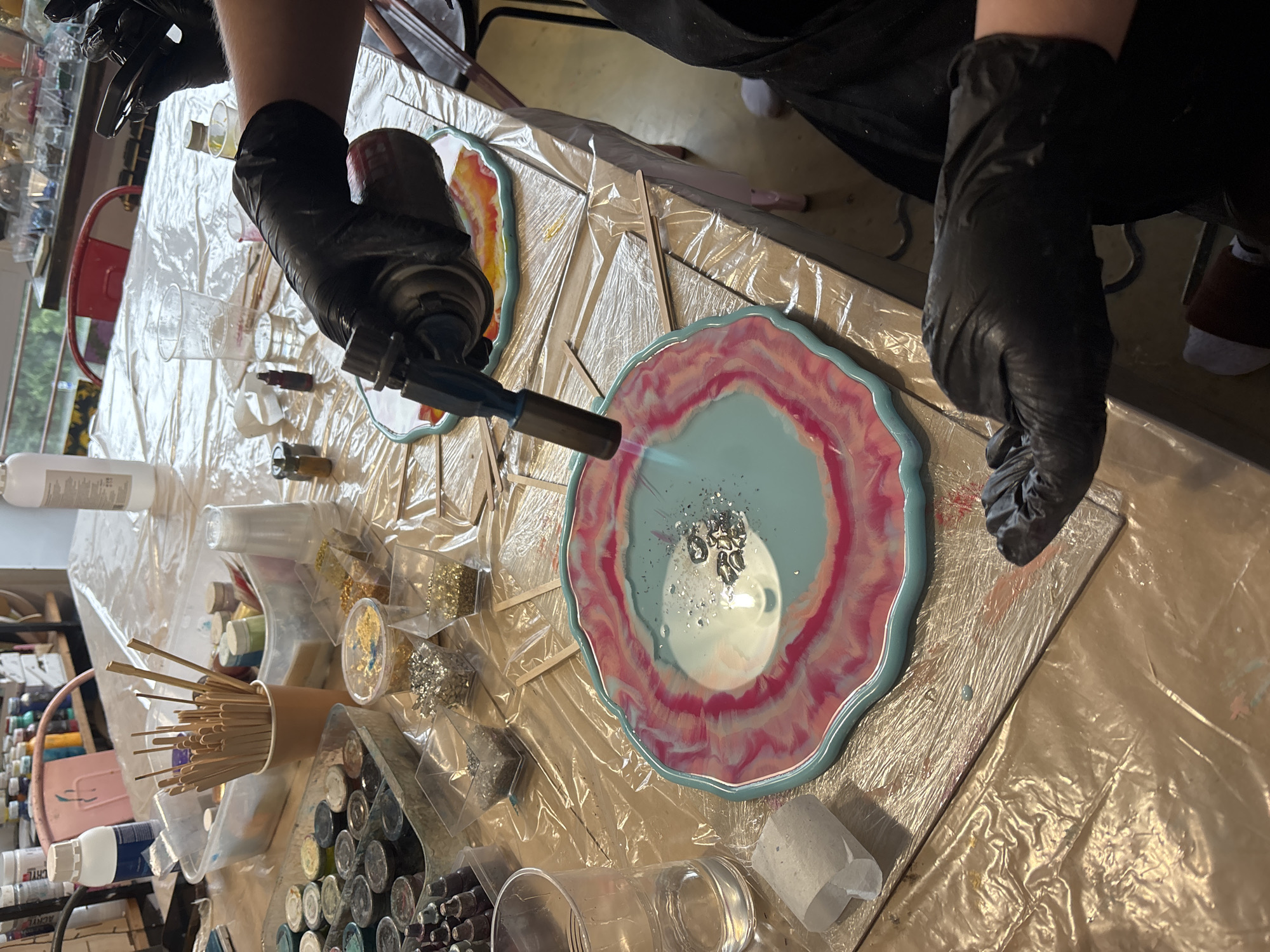 In an indoor art studio, likely a home workshop in Naberezhnye Chelny, Russia, a person wearing black nitrile gloves is using a butane torch to heat a teal and pink resin art piece. The art piece, shaped like a geode, is on a table covered with a clear plastic sheeting. Scattered around the table are various art supplies including small containers of glitter in silver and gold, tubes of colored resin dyes, stirring sticks, and plastic cups. In the background, shelves are stocked with bottles of art materials. The overall scene suggests a creative process involving resin casting and decoration.