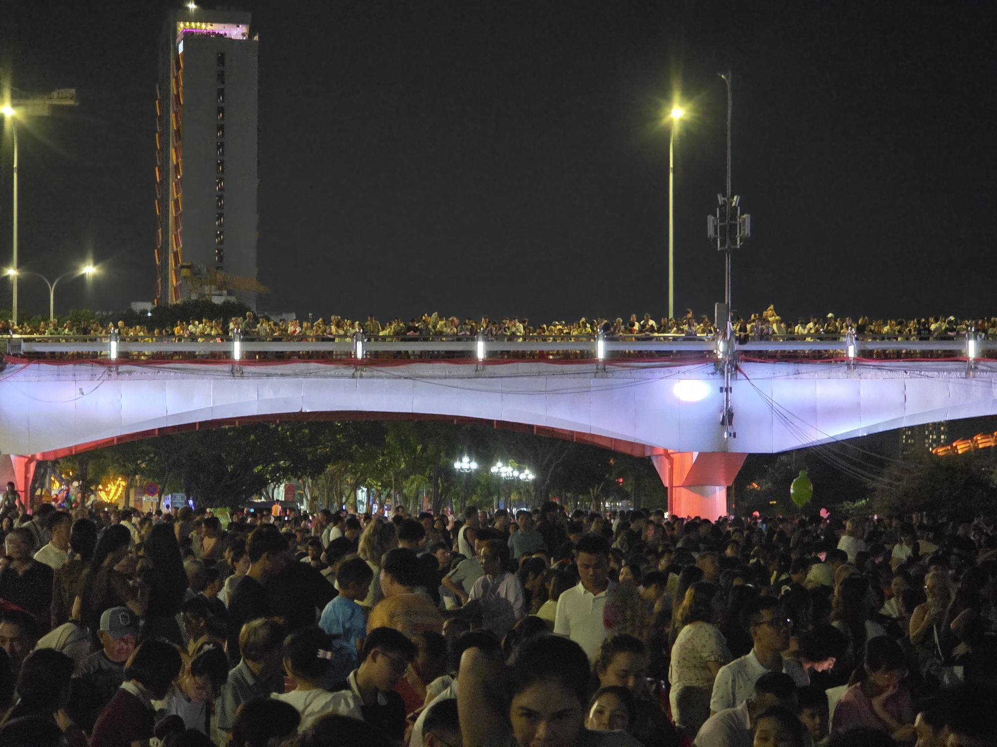 This image depicts a large crowd of people gathered in the city of Đà Nẵng, Vietnam. In the foreground, a dense group of people, including children, is densely packed in front of a white bridge, which is lit with red and white lights. A crowd of people also stands on the bridge. To the left of the bridge is a tall, modern building with lights. Light posts are seen around the edge of the bridge and city. The overall scene appears to be captured at night.