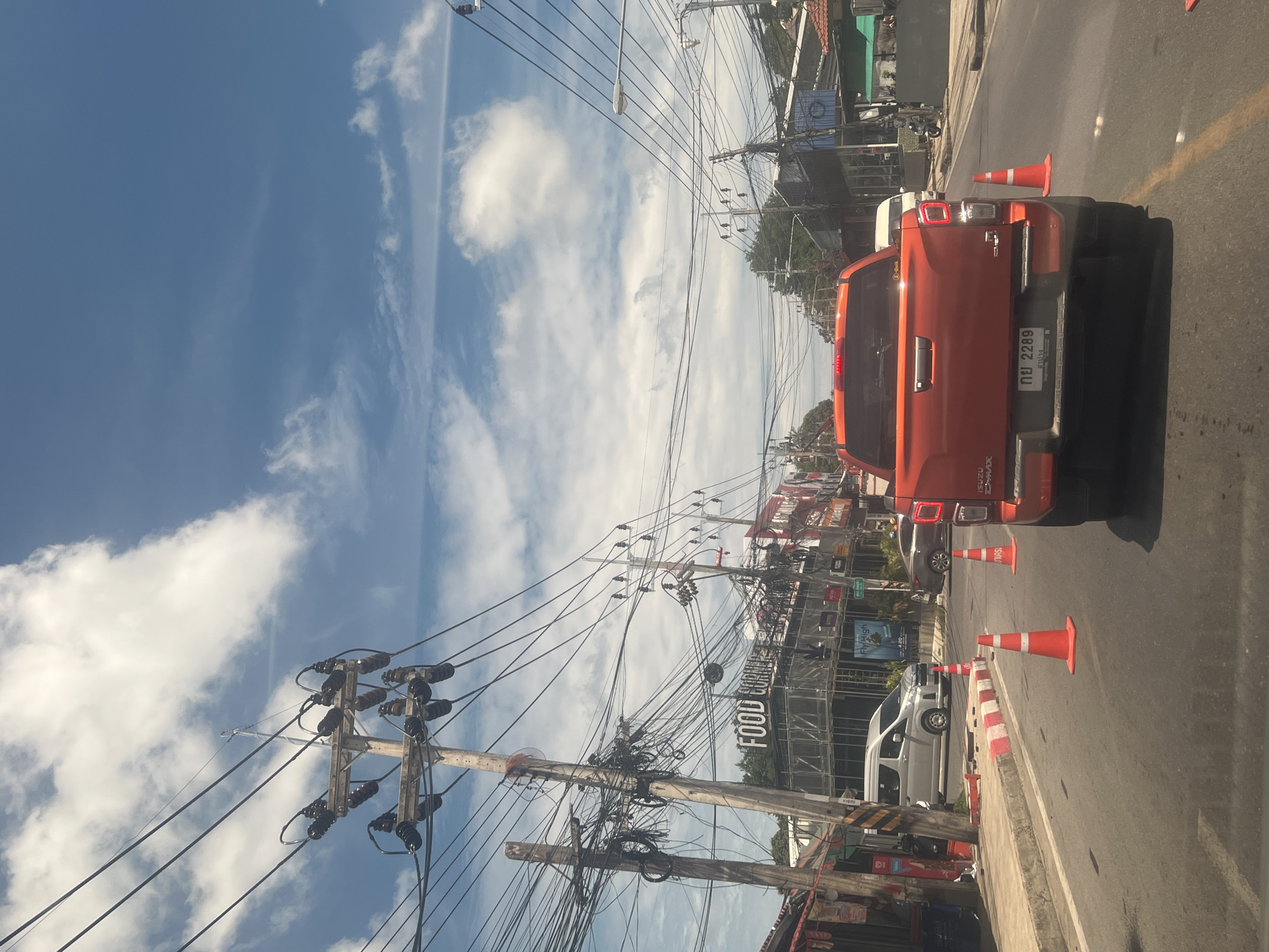 An orange pickup truck with a license plate that reads "กย 2289 ลำปาง" is stopped on a road, viewed from behind. The truck is in the right lane. Ahead of the truck are traffic cones on the road. To the left of the truck, a silver van is stopped in the left lane. The scene appears to be a street in a city with buildings and businesses lining the road. Overhead, a dense network of electrical wires crisscrosses the sky, supported by numerous utility poles. The sky is blue with scattered white clouds. The buildings visible include one with a sign that says "FOOD CORNER". The time of day appears to be daytime, with bright sunlight. This scene is consistent with a busy street in Ko Samui District, Thailand, likely a commercial area.
