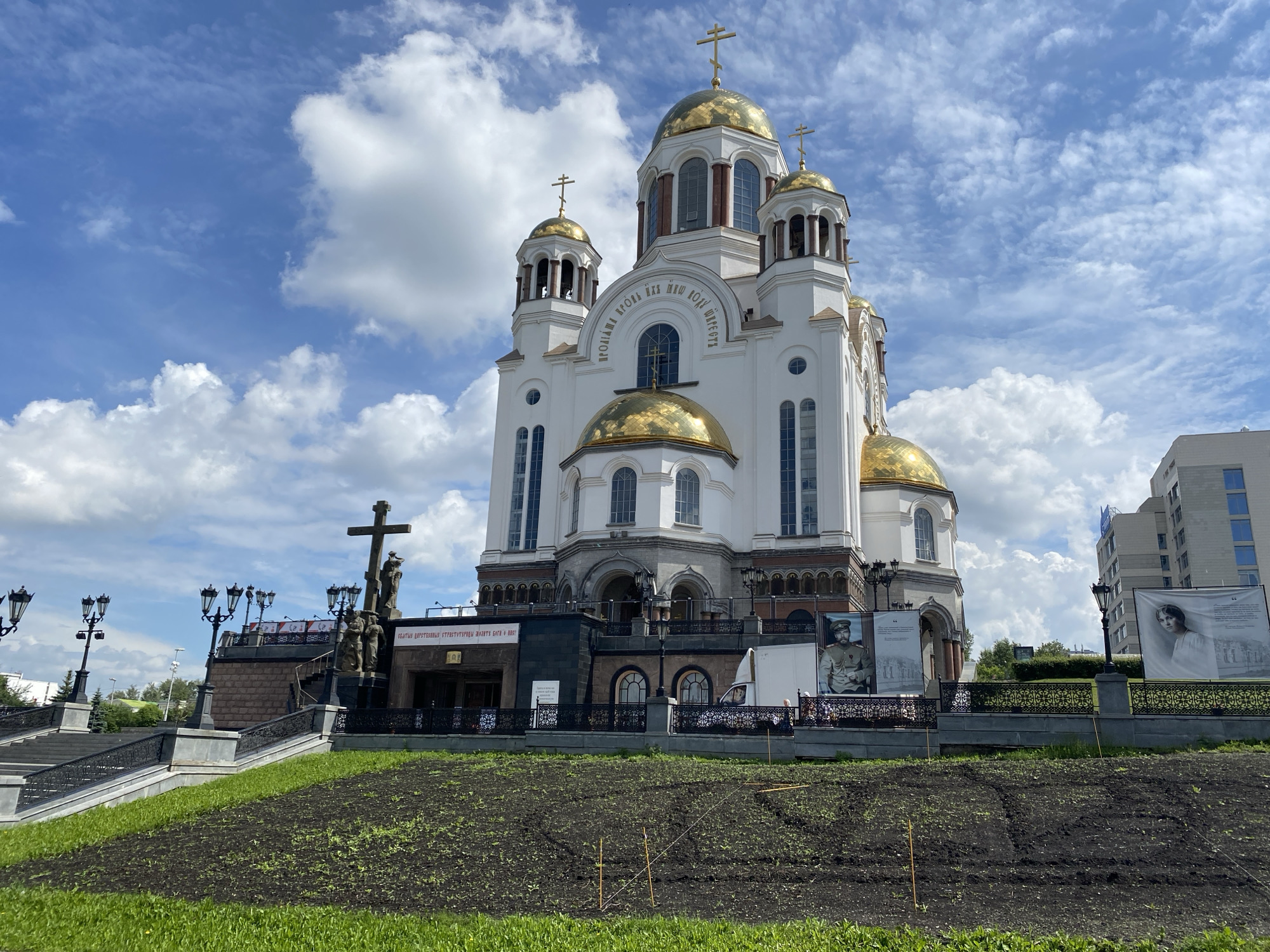 The image shows the Church on Blood in the city of Yekaterinburg, Russia, under a cloudy blue sky. The church is a large, white building with golden domes and crosses. In front of the church, there is a large monument with a cross and several figures. There are also banners displayed on the fence in front of the church, featuring portraits of historical figures. In the foreground, there is a grassy area with freshly tilled soil, suggesting recent landscaping or planting. Modern buildings can be seen in the background.