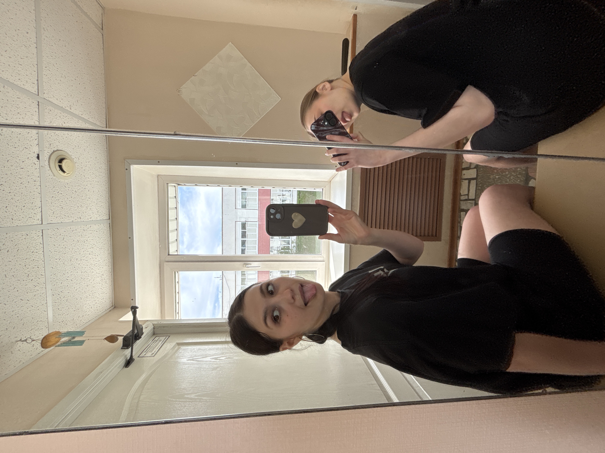 A young woman takes a mirror selfie in what appears to be a residential building or apartment complex. The reflection shows the woman, wearing a dark t-shirt and shorts, holding a smartphone with a heart-shaped design on the back. She is sticking her tongue out in a playful manner. The mirror also reflects another person above her, partially visible, holding another phone.
The environment includes a ceiling with a white tiled pattern, a window showing an apartment building across the street, and a door with visible hardware. Based on the architectural style of the buildings visible through the window, and the assumption that this photo was taken in Russia, the location is possibly in or around an apartment building in Chaikovsky, Russia, considering the user's prompt.
