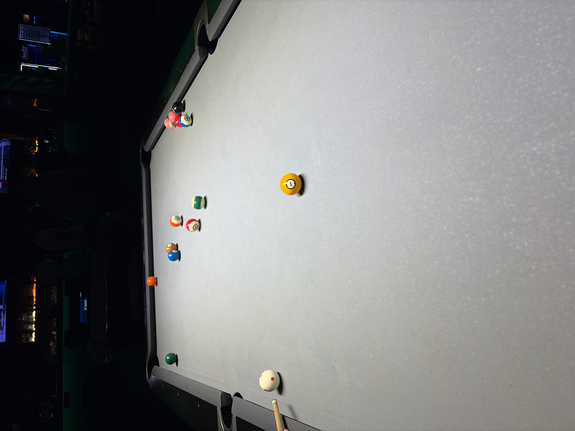 A pool table is visible in a dimly lit establishment, likely a bar or pub. The table is covered in a light gray felt and several pool balls are scattered across it. A cue stick is positioned on the left side of the table, suggesting a game is in progress or has recently concluded. The background is dark and out of focus, with hints of bar shelves, bottles, and lighting visible. There are no people clearly depicted in the foreground, but shadowy figures can be seen in the background.