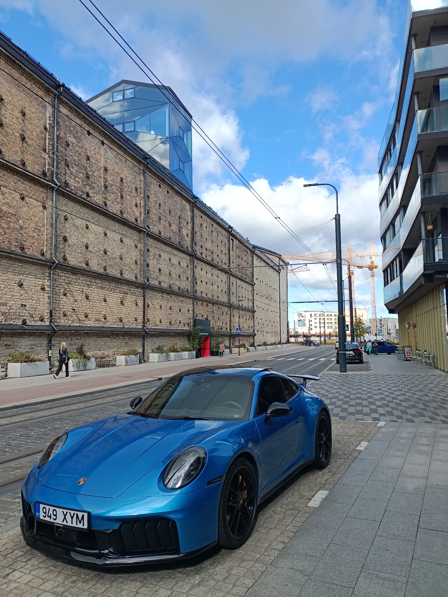 In Tallinn, Estonia, a bright blue Porsche 911 GT3 is parked on a cobblestone street in front of a modern building with glass balconies. To the left, an older brick building with distinctive circular protrusions lines the street. A woman in black clothing walks on the sidewalk beside the brick building. In the background, yellow construction cranes are visible against a cloudy blue sky. The scene suggests an urban environment with a mix of historical and contemporary architecture, hinting at ongoing development or renovation.