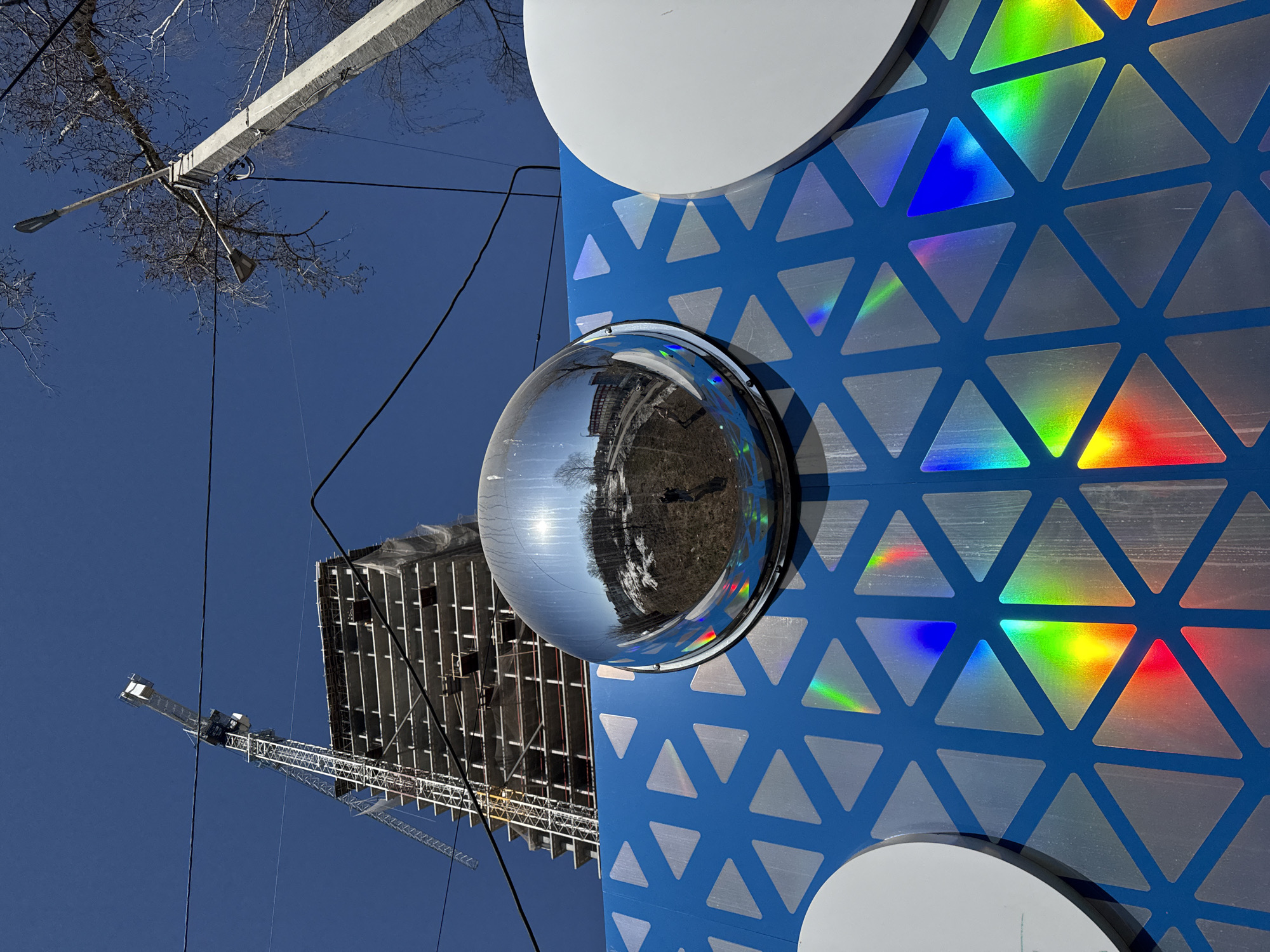 The image, taken from a low angle, features a modern art installation against a clear blue sky. The installation is a blue structure adorned with iridescent triangles and circular white elements. A chrome sphere is attached to the blue surface, reflecting a building under construction, barren trees, and a construction crane. In the background, utility poles and bare tree branches are visible against the sky. The scene captures the juxtaposition of art and construction in an urban environment, located in Izhevsk, Russia.