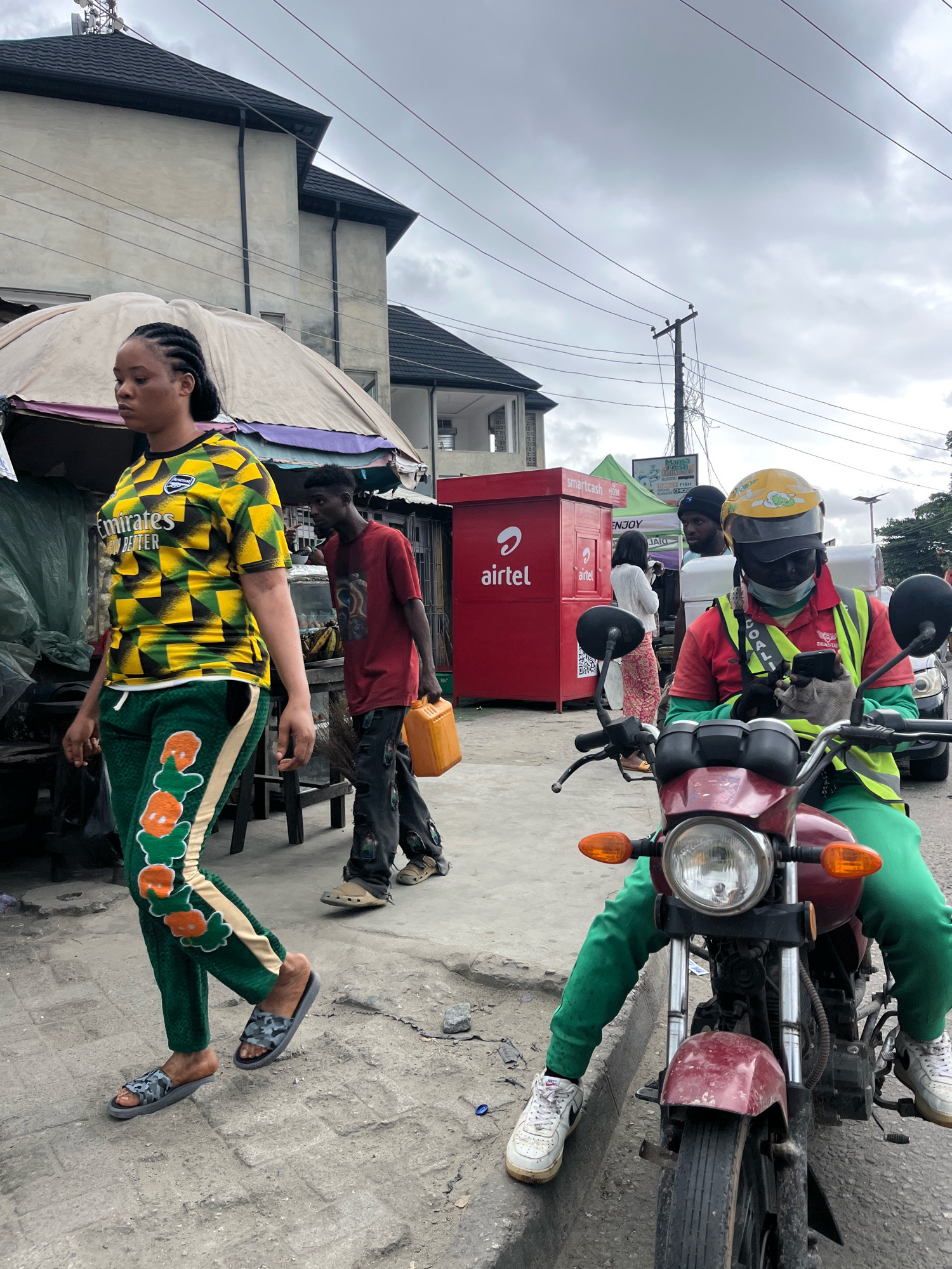 In Ado, Nigeria, under an overcast sky, a scene unfolds on a paved street lined with buildings and temporary stalls. In the foreground, a person on a maroon motorcycle, wearing a yellow helmet, a red shirt, a neon green vest, and green pants, looks at their phone. They are wearing gray gloves and white sneakers. To their left, a woman with braided hair walks away from the camera, wearing a yellow and green patterned shirt with "Emirates" branding and green pants adorned with orange and white floral designs. She is wearing camouflage flip-flops. Behind her, a man in a red t-shirt and dark pants carries an orange plastic container. Further in the background, a red Airtel kiosk is visible, along with other people and structures. The environment appears to be a bustling urban area with visible power lines crisscrossing the sky.