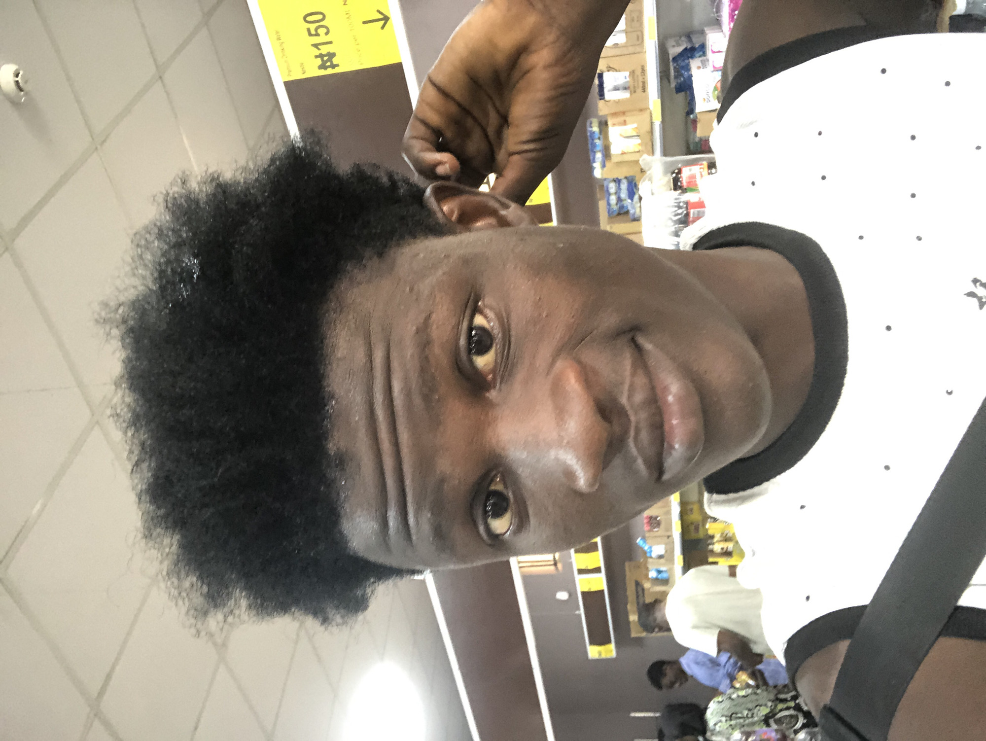 In the city of Ado, Nigeria, a young Black male takes a selfie in what appears to be a retail store. He has dark, curly hair and is looking at the camera with a slight smile, his right hand touching his ear. He is wearing a white tank top with black trim and small black dots. In the background, shelves stocked with various products are visible, along with other shoppers. A yellow sign with the currency symbol "₦" and the number "150" is also present, indicating pricing. The ceiling lights suggest an indoor environment.