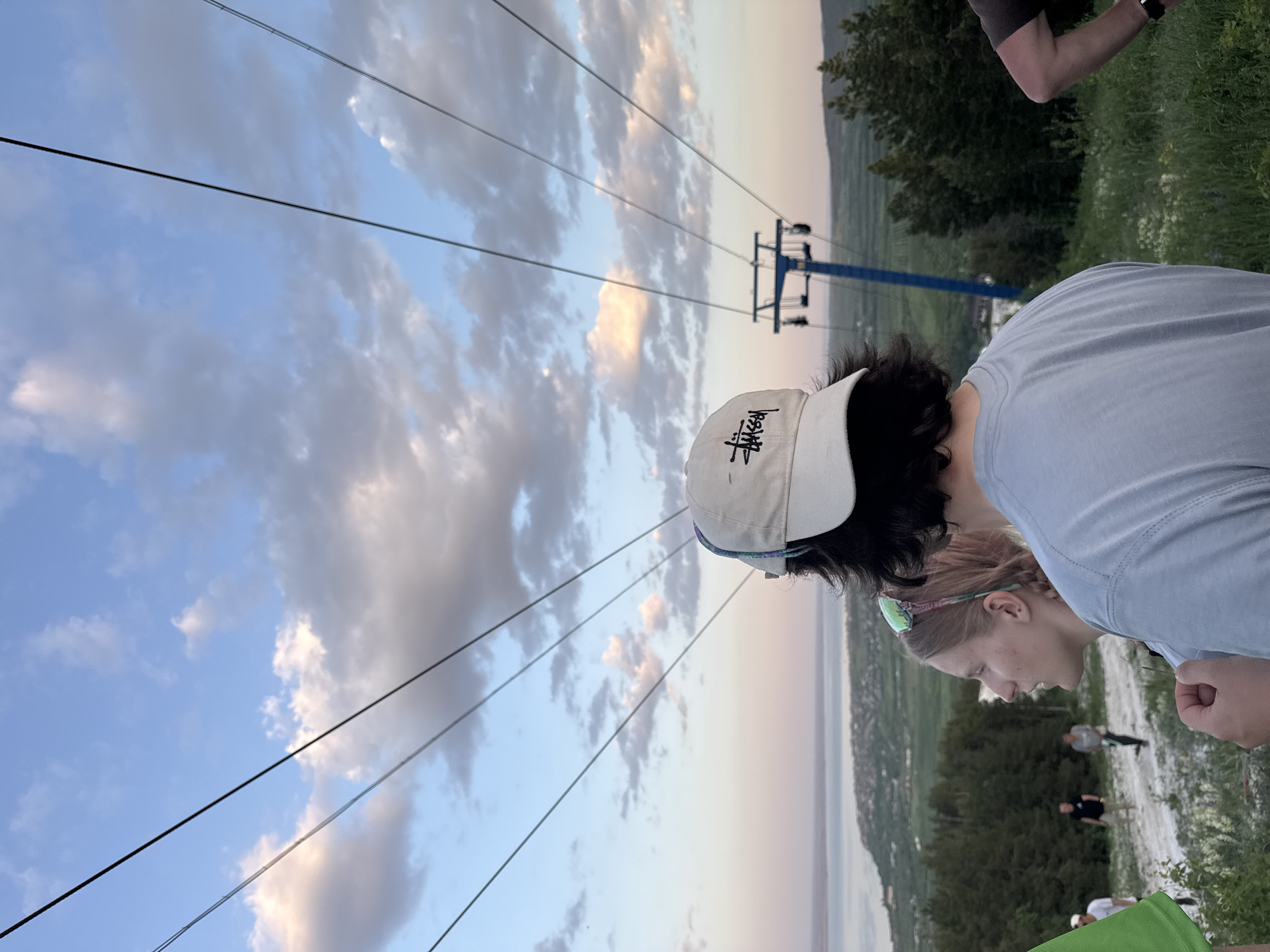 Two people are standing outdoors on a grassy area. A woman with fair skin and blonde hair pulled back is visible in profile. A person is wearing a light-colored cap with black lettering. In the background, there is a blue ski lift structure. A lake is partially visible. The sky is cloudy with hues of blue, white, and pink. Power lines run diagonally across the image. Other individuals can be seen indistinctly in the middle ground, likely on a path. The location is Khvalynsk, Russia.