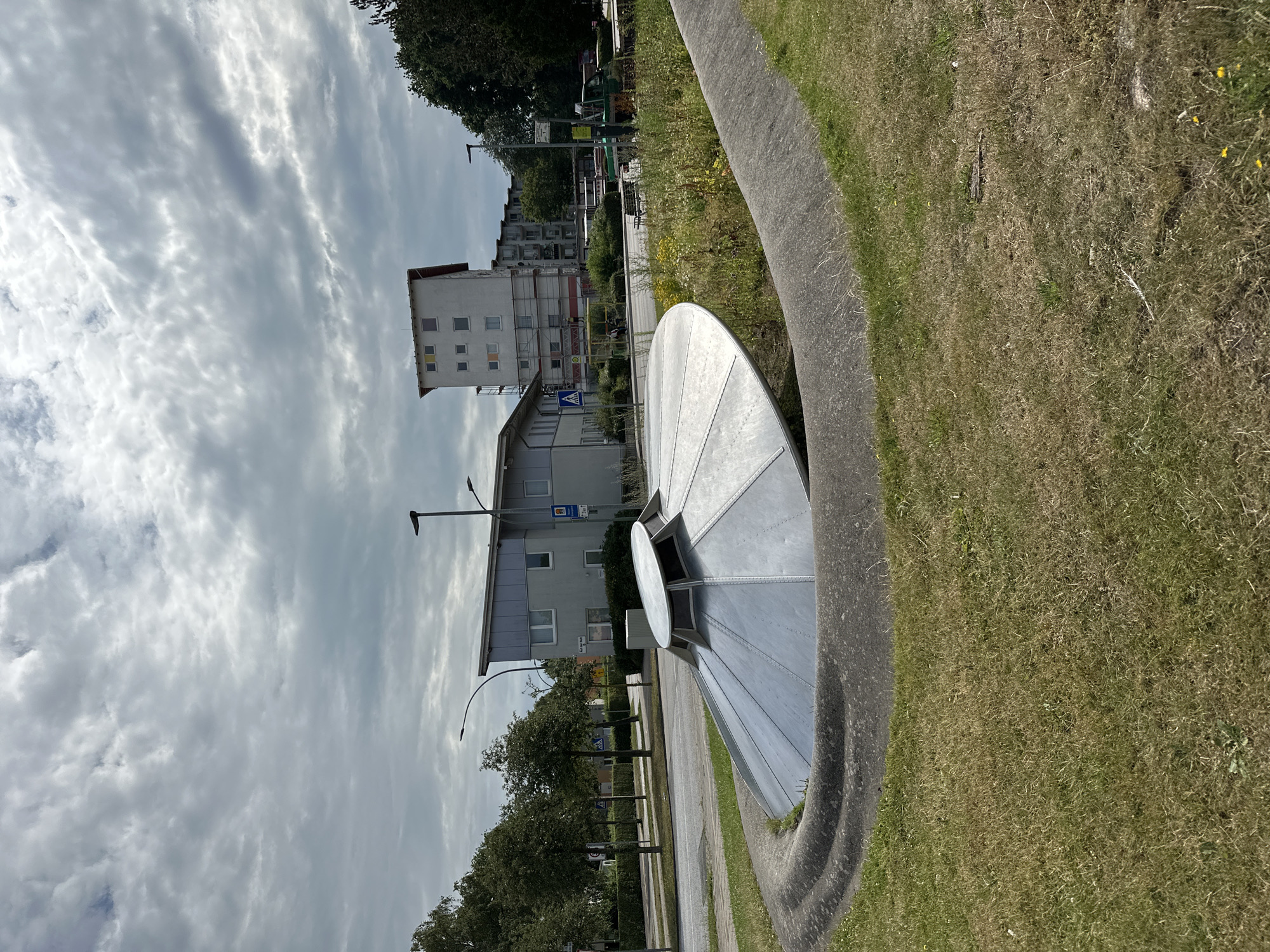 The image shows a large, metallic, saucer-shaped sculpture resembling a UFO, partially embedded in the ground and surrounded by a concrete border. The sculpture is silver and appears to be constructed from riveted panels. It is situated in a grassy area with some wildflowers and weeds growing around it.
In the background, a street is visible with trees lining one side. Across the street, there are several buildings, including a light-colored, multi-story apartment building with red trim and a lower, gray building with windows. Streetlights and traffic signs are also present. The sky is overcast with textured clouds.
This sculpture is located in Bremerhaven, Germany, specifically in the "Alte Bürger" district, at the intersection of "Bürgermeister-Smidt-Straße" and "Steinstraße." It is a public art installation and a recognizable landmark in the area. There are no people or animals clearly visible in the image. The overall scene depicts an outdoor urban environment with a prominent art piece.