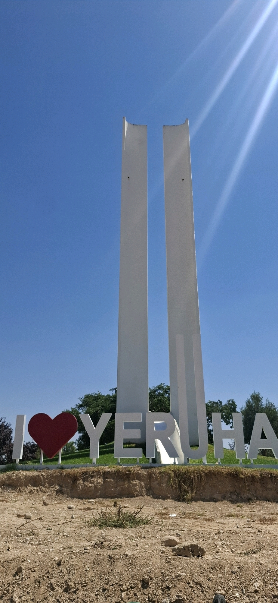 The image shows the "I Love Yerocham" sign in the city of Yerocham, Israel. The sign features large, white, three-dimensional letters spelling out "YERUHAM," with a prominent red heart symbol in place of the word "LOVE." Behind the sign, two tall, slender white pillars stand side-by-side, reaching towards a clear blue sky. Sunlight beams down from the upper right corner of the frame, creating bright rays across the sky. The sign is situated on a grassy area, with the foreground showing a rough, dirt-covered ground with scattered rocks and sparse vegetation. The overall scene depicts a sunny day in an outdoor public space.