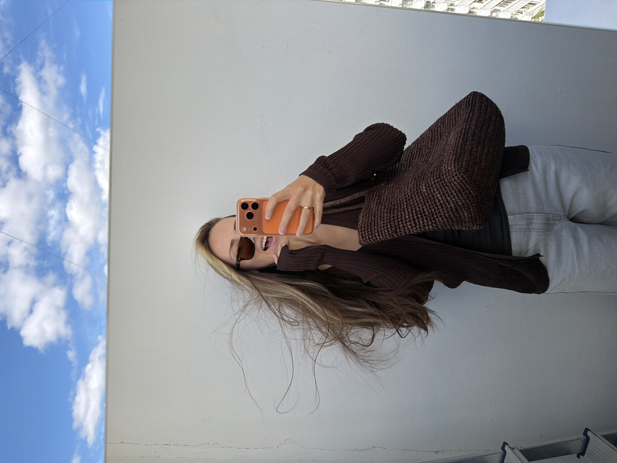 In Khimki, Russia, a young woman with long, windswept blonde hair stands on a rooftop, holding up a smartphone in her right hand and taking a selfie. She is wearing sunglasses, a dark brown cardigan over a darker top, and light grey pants. A dark brown woven tote bag hangs from her left arm. Behind her is a plain white wall, and above that, a bright blue sky with scattered white clouds. To the right of the wall, the reflection in a mirror reveals a portion of a building with multiple windows. The overall scene suggests a casual, outdoor setting on a sunny day.
