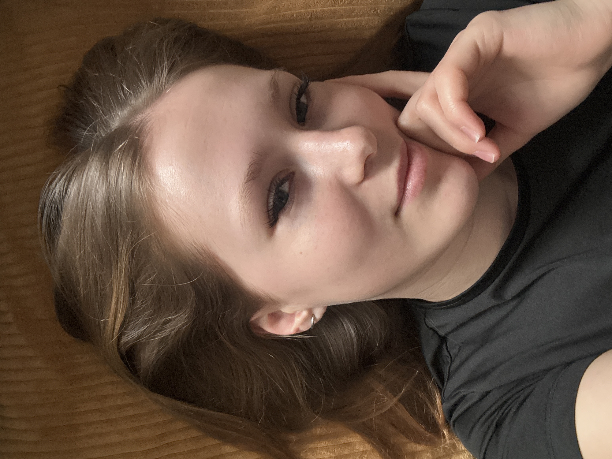 A close-up, slightly angled portrait of a young woman with light skin and brown hair. She is lying down on what appears to be a textured, brown surface, possibly a blanket or bedspread. The woman is wearing a dark-colored shirt. Her right hand is gently touching her face near her cheek. She is looking directly at the camera with a slight smile. The location is not discernible from the provided image. Therefore, there is no indication that the image was taken in Chaikovsky, Russia.