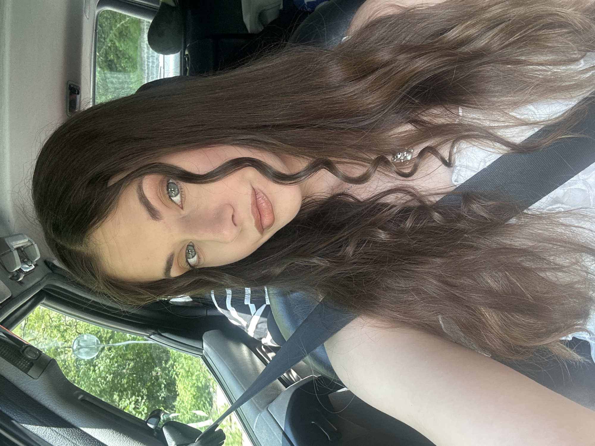A woman with long, dark brown hair and green eyes is sitting in the driver's seat of a vehicle. She is wearing a black and white striped shirt and a seatbelt. The interior of the vehicle is visible, with the dashboard and side window in view. Outside the window, trees and foliage are visible, suggesting a possibly rural or suburban environment. The city is Unknown, Unknown.