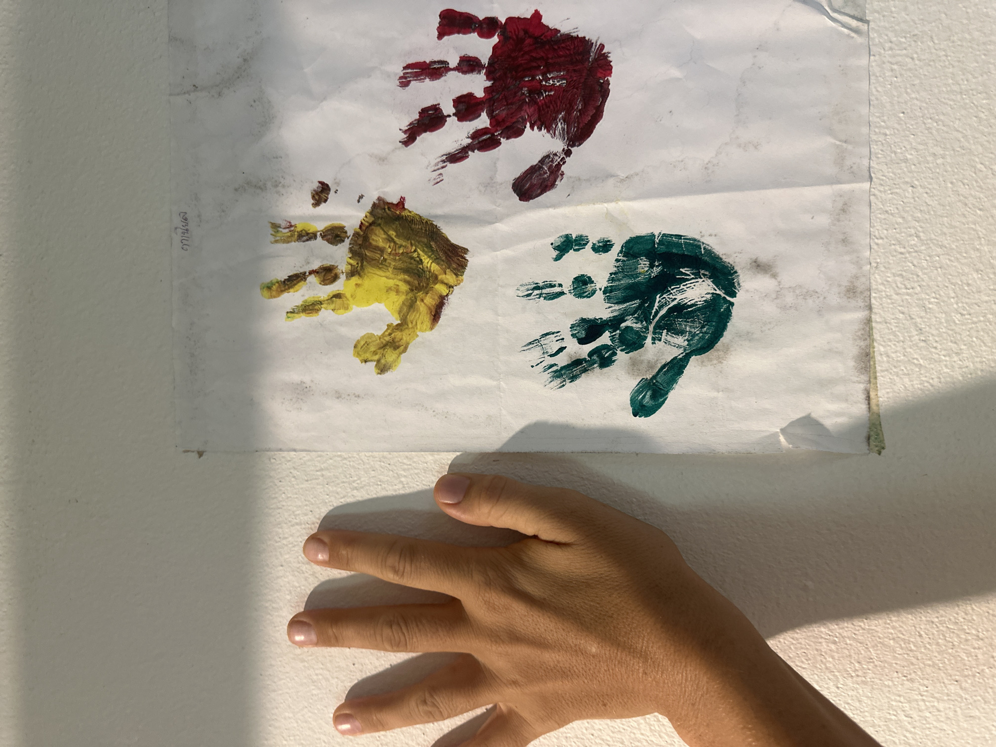 A close-up, overhead shot shows a person's left hand with fingers slightly spread, touching a white, textured wall. To the right of the hand, a piece of paper is taped to the wall, featuring three colorful handprints: yellow, green, and dark red. The yellow handprint is at the top, the green handprint is at the bottom left, and the red handprint is at the top right. Above the handprints, faint writing in what appears to be Thai script is visible on the paper. The lighting suggests an outdoor environment, with shadows cast by the hand onto the wall. The scene is located in ตำบลเกาะพะงัน, Thailand.