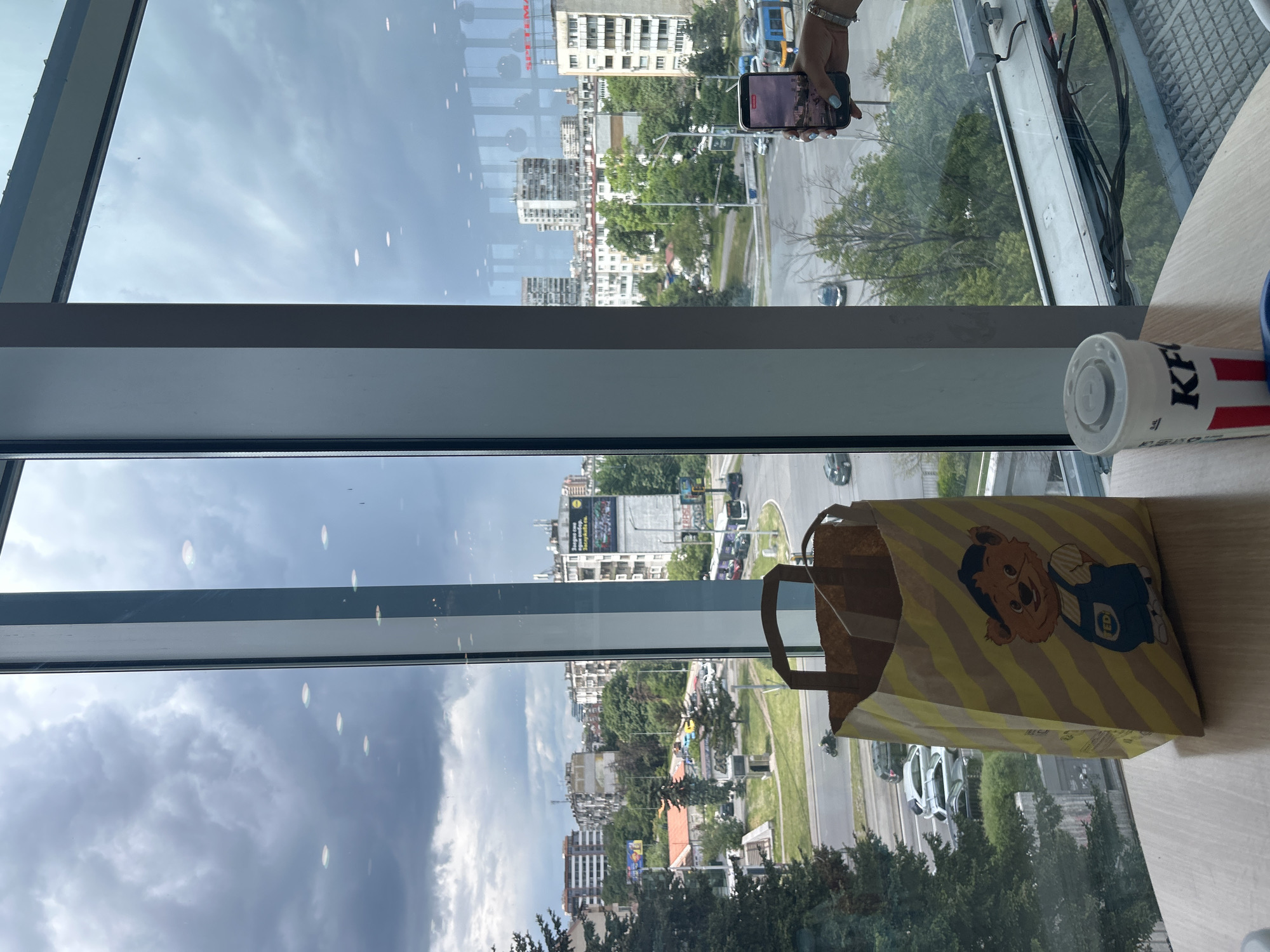 A person is holding a smartphone facing towards a window in a high-rise building. The smartphone is pointed toward the cityscape outside the window. The view outside the window includes various buildings, trees, and streets with moving vehicles. Part of a table is visible with a KFC drink and a yellow and brown striped bag.