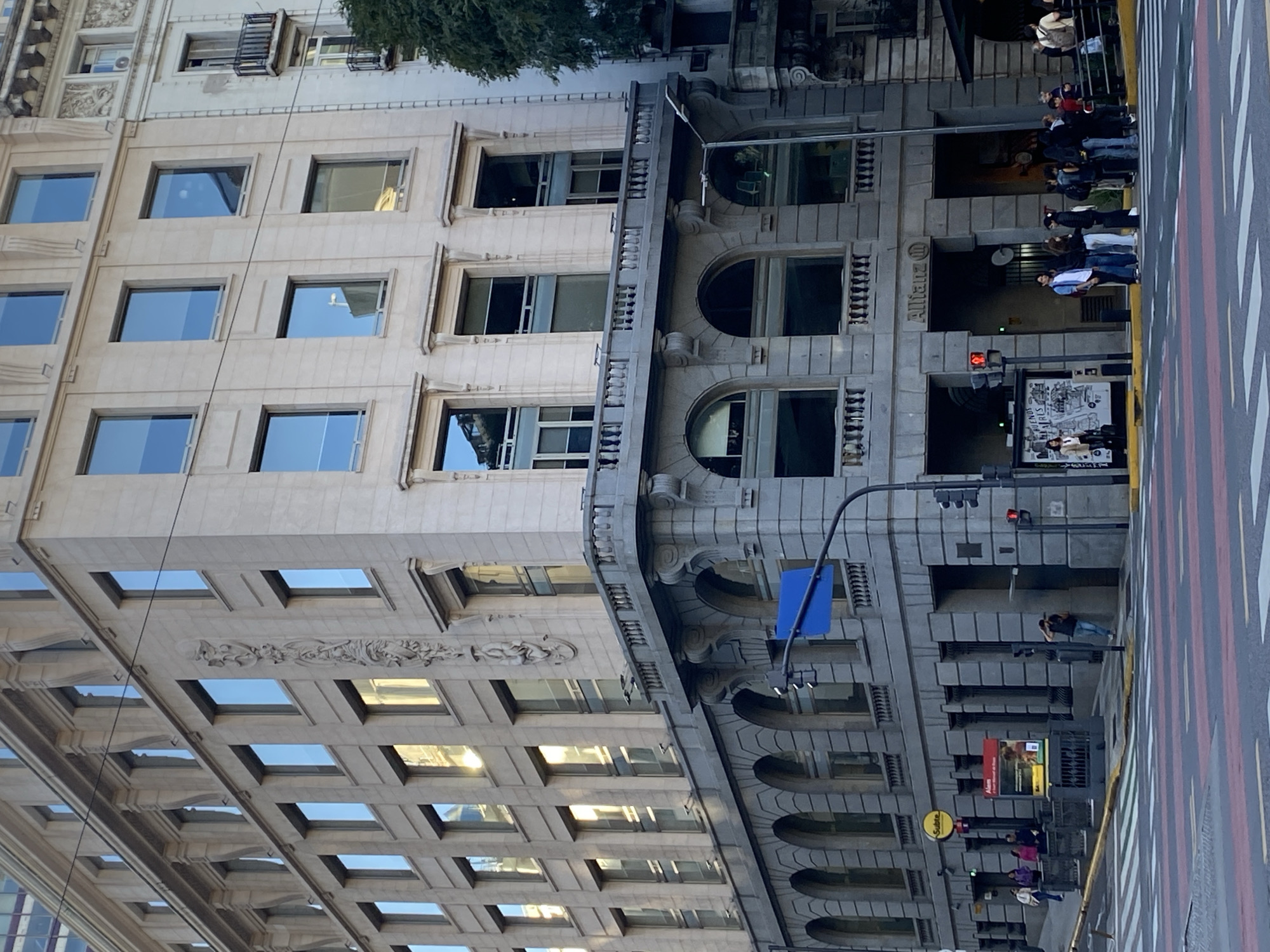 The image depicts a street scene in Buenos Aires, Argentina. The main focus is on two adjacent buildings. One building has a light-colored facade with rectangular windows in a grid pattern, reflecting sunlight. The other building has a darker, gray facade with arched windows and ornate architectural details. The "Allianz" sign is visible on the darker building. A group of people are standing on the sidewalk, waiting to cross the street. A crosswalk with colored stripes is visible on the road. There is a traffic light with a red signal. A blue street sign is also visible. A street pole with a small yellow sign is also present. There is also a poster on the side of the building. One individual is standing alone in the crosswalk.