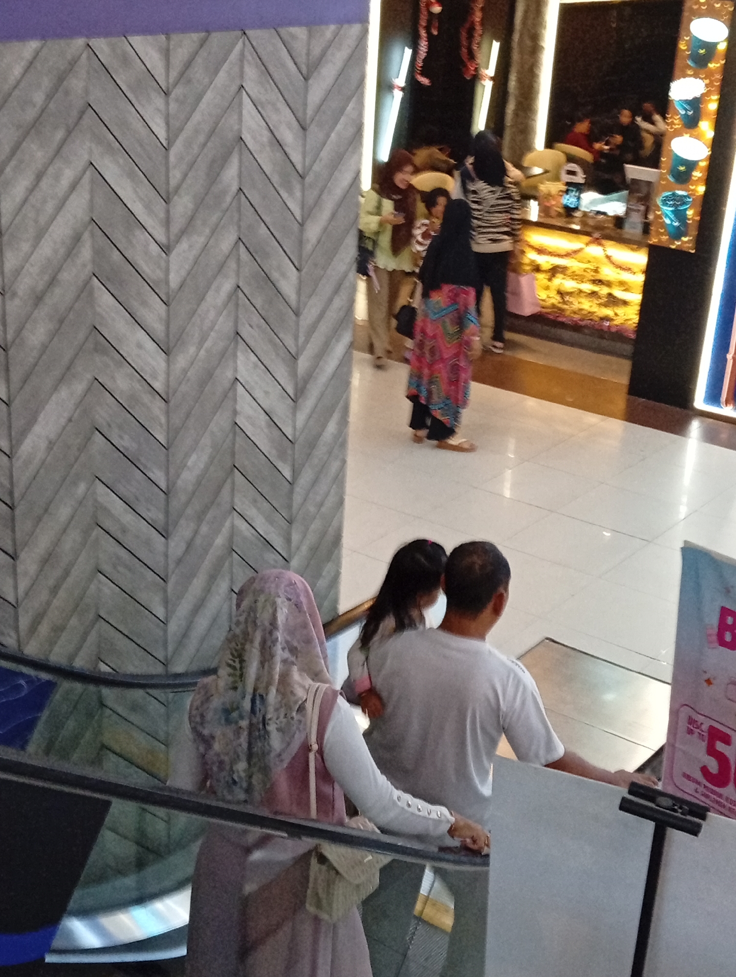 The image shows an indoor scene, likely a shopping mall in Sidoarjo, Indonesia. In the foreground, two people, an adult male and a young female, are seen from behind as they stand near an escalator. The male wears a white t-shirt, and the female has long dark hair. To their left and slightly in the background, a woman in a headscarf and a long pink dress is visible on the escalator, also seen from behind. Further back, a brightly lit counter area is observed with several people interacting. The walls feature a distinct grey chevron pattern on the left side of the image, and the floor is tiled. A large poster with pink and white text is visible on the right side of the frame.