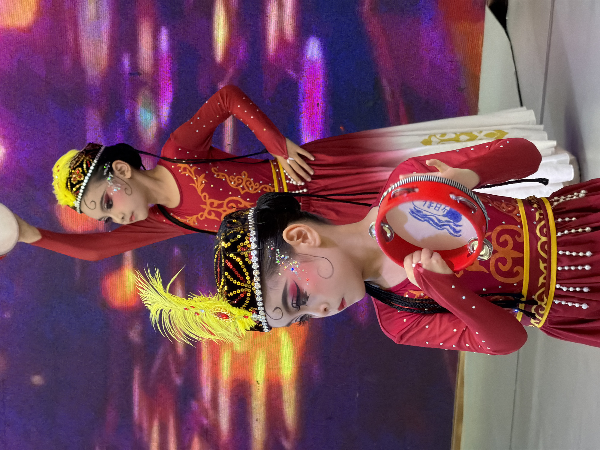 Two young girls in traditional red and gold costumes are performing on a stage. The girl in the foreground, on the left, wears a black, sequined hat adorned with a large yellow feather. She has heavy eye makeup with glitter accents on her cheeks and is holding a red tambourine. The girl in the background, on the right, also wears a similarly decorated hat with yellow plumage and is holding a tambourine above her head. They are positioned in front of a vibrant, blurred background of purple, pink, and orange hues, suggestive of a stage curtain or backdrop. The setting appears to be an indoor performance space, possibly a school or community center, in the Jiulongpo District of China.