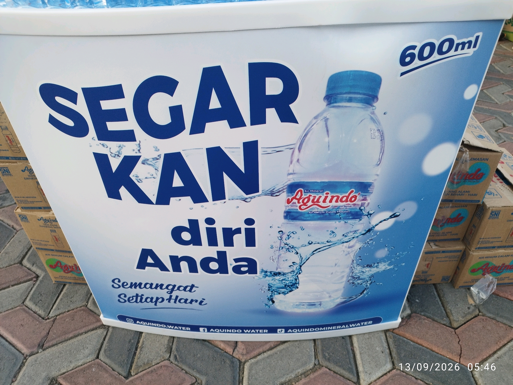 The image shows a white display board with blue text and a picture of a bottle of Aquindo mineral water. The text on the board reads "SEGAR KAN diri Anda" which translates to "REFRESH YOURSELF". Below this, it says "Semangat SetiapHari" meaning "Spirit EveryDay". The Aquindo logo is visible on the water bottle. The display board is placed on a patterned brick surface, likely a sidewalk or pedestrian area. In the background, there are stacked cardboard boxes, some with the Aquindo logo, suggesting a retail or promotional setting. The timestamp at the bottom right indicates the photo was taken on September 13, 2026, at 05:46. The overall scene is outdoors, with natural lighting. While the image does not definitively place the location within Mojokerto, Indonesia, the presence of Indonesian language text on the display and product packaging strongly suggests it.
