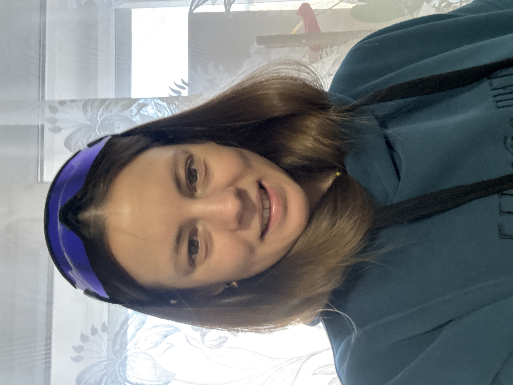 A woman is pictured indoors, likely in a residential setting, given the floral curtain and window. She has brown hair, fair skin, and is wearing ski goggles pushed up on her forehead. She is also wearing a teal hooded sweatshirt. The location is not explicitly identifiable as Chaikovsky, Russia from the image alone.