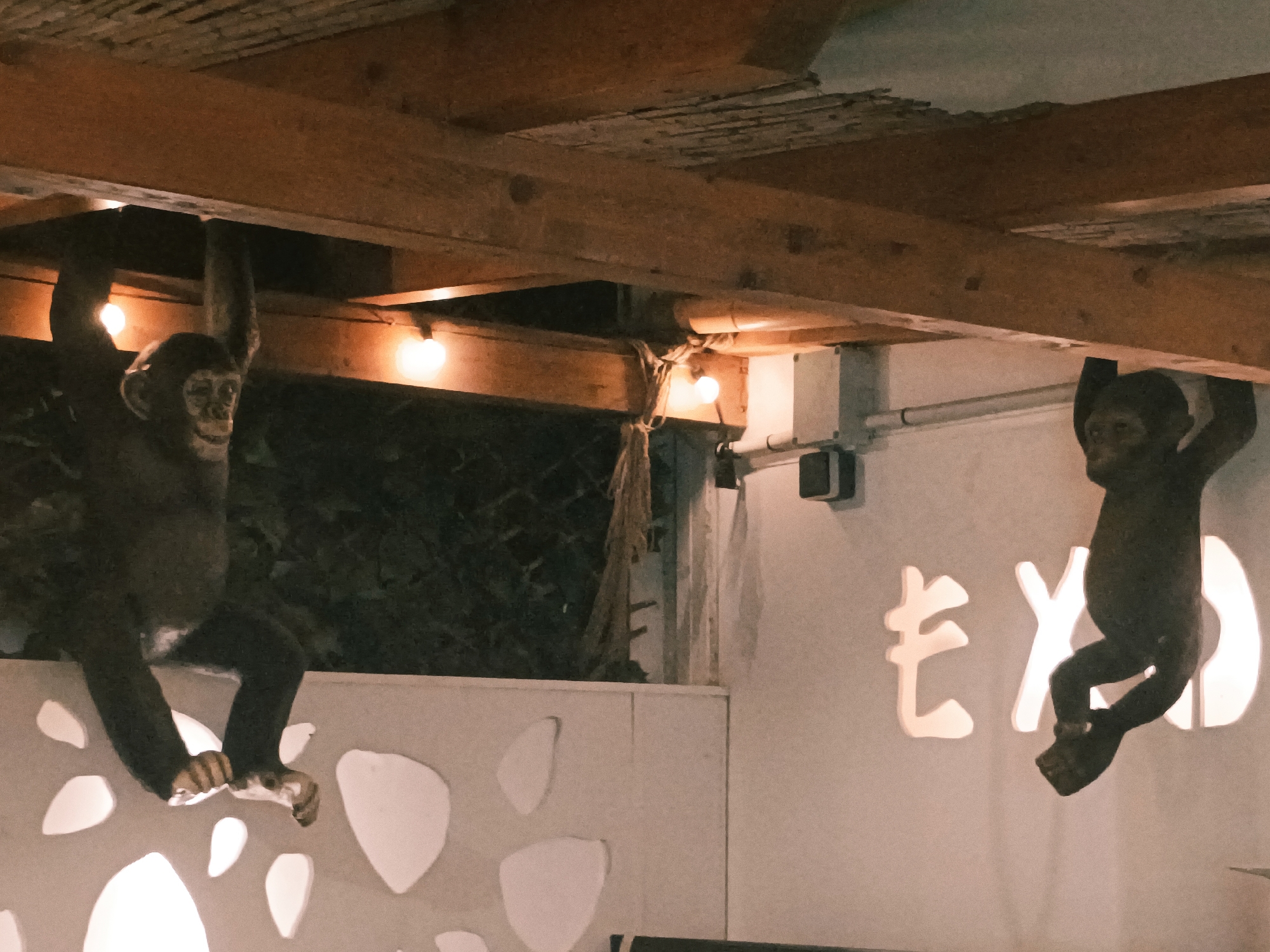 In San Gabriel, Spain, two monkey figurines are suspended from wooden beams above a white wall. The figurine on the left is larger and appears to be holding onto the beam with both hands, with its legs dangling. The figurine on the right is smaller, hanging by its hands from another beam, with its legs bent. Behind the figurines, on the white wall, a glowing Japanese character is visible. The scene is lit by several lightbulbs, creating a warm and atmospheric environment. The ceiling is made of rough wooden planks.