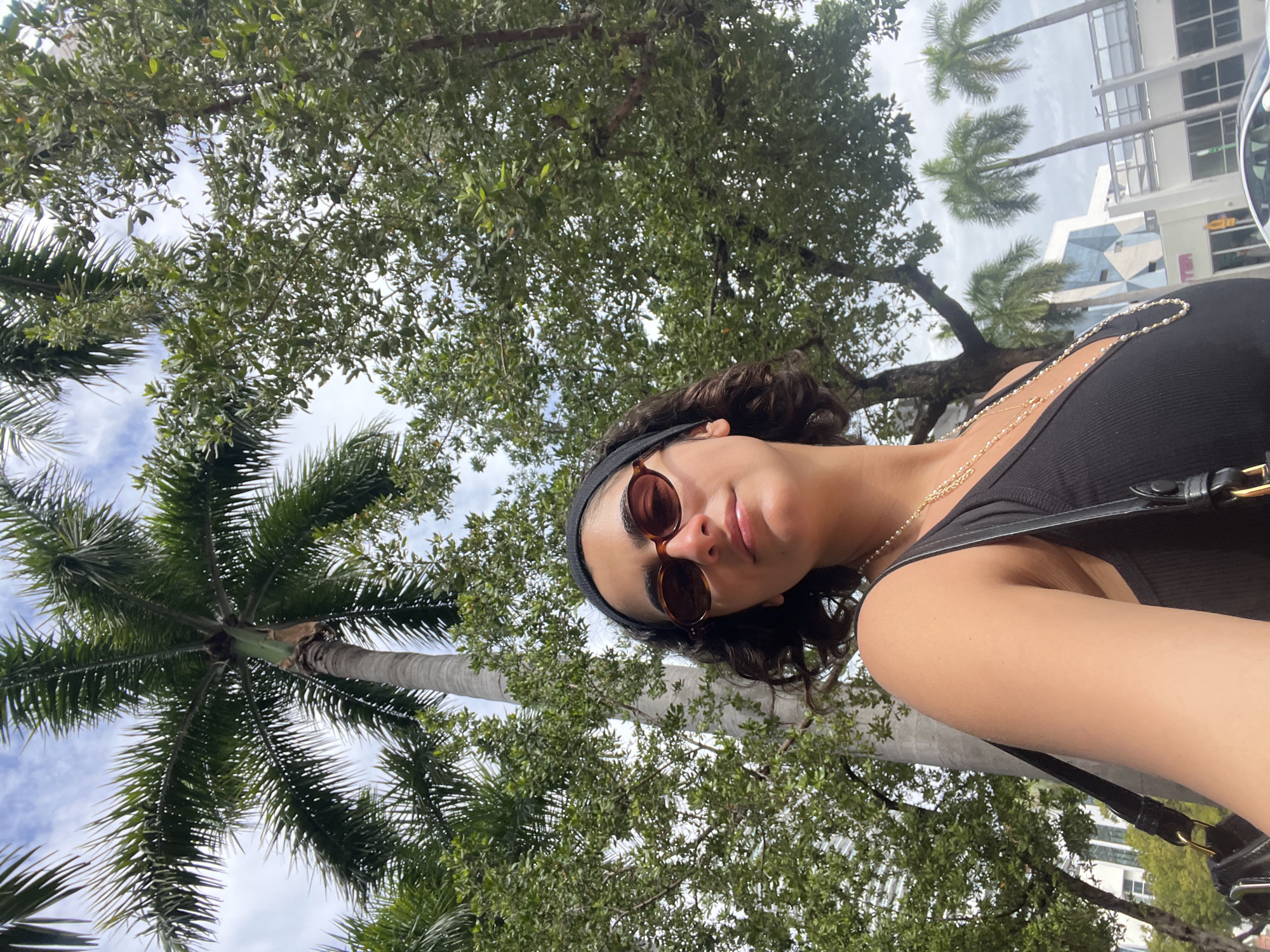 A medium-angle shot shows a female wearing dark-tinted sunglasses and a black headband, looking downward. She is wearing a black top with a gold necklace. She also has a black purse over her shoulder. The background features a canopy of green trees, including palm trees. In the upper right of the frame, the top of a tall building can be seen. Based on the architecture, the trees, and the sky, the location is possibly Miami Beach, United States of America. The image is taken outdoors during the daytime.