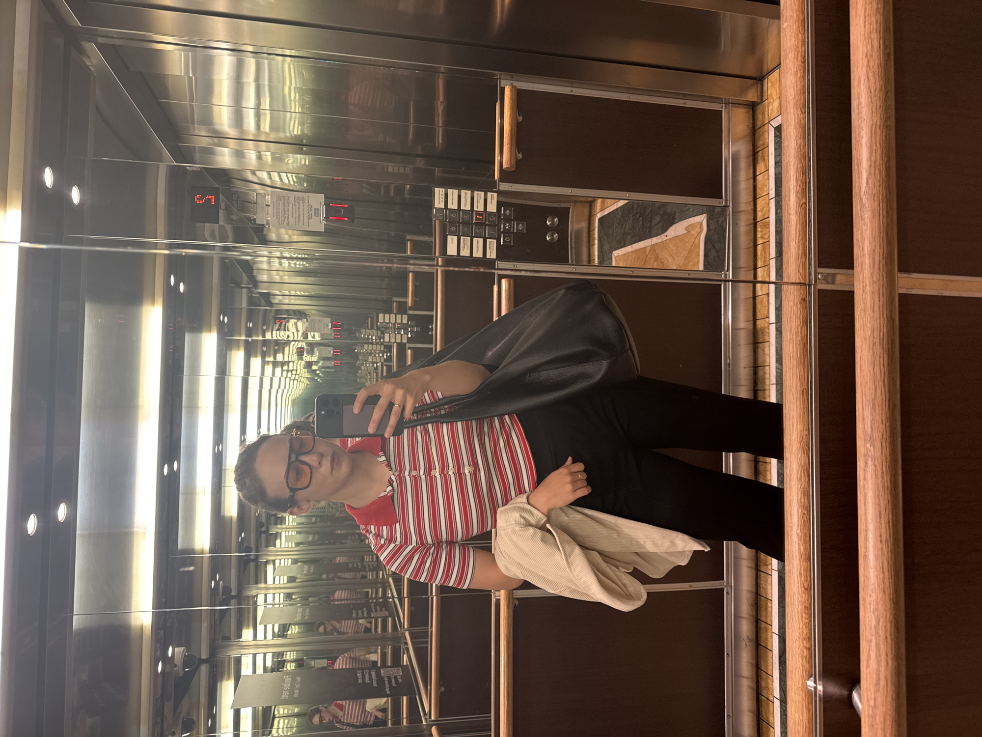 A woman with brown hair pulled back, wearing large orange-tinted glasses, a red and white striped polo shirt, and black pants, stands in an elevator. She is holding a beige jacket over her left arm and a large black handbag in her right hand. She is taking a selfie with a smartphone in her right hand. The elevator has mirrored walls, stainless steel accents, and wooden handrails. The floor is patterned with marble and tile. An elevator panel with buttons and a digital floor indicator showing "5" is visible on the right wall. The scene is well-lit, reflecting the interior of the elevator. The location is identified as an elevator in Riga, Latvia, based on the visible signage, which includes text in Latvian.