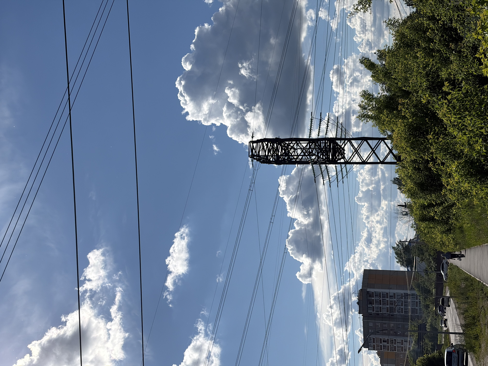 The image shows a view in Chaikovsky, Russia. An electrical transmission tower is centered in the frame. Power lines stretch across the bright, blue sky filled with white clouds. A tree line flanks the right side of the image with an apartment building visible below. Cars and pedestrians are on the street.